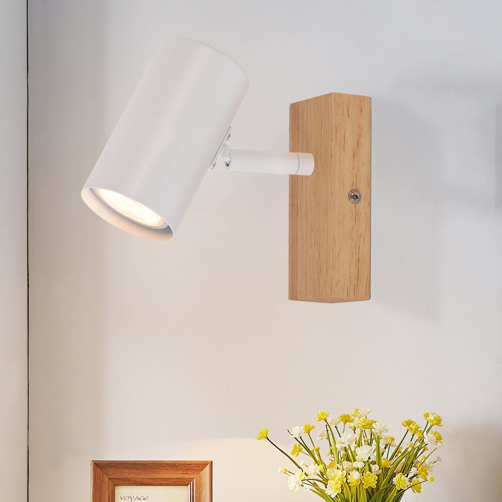 Weißer Strahler auf Holzbasis, beleuchtet Wand, kleine Blumenanordnung darunter.