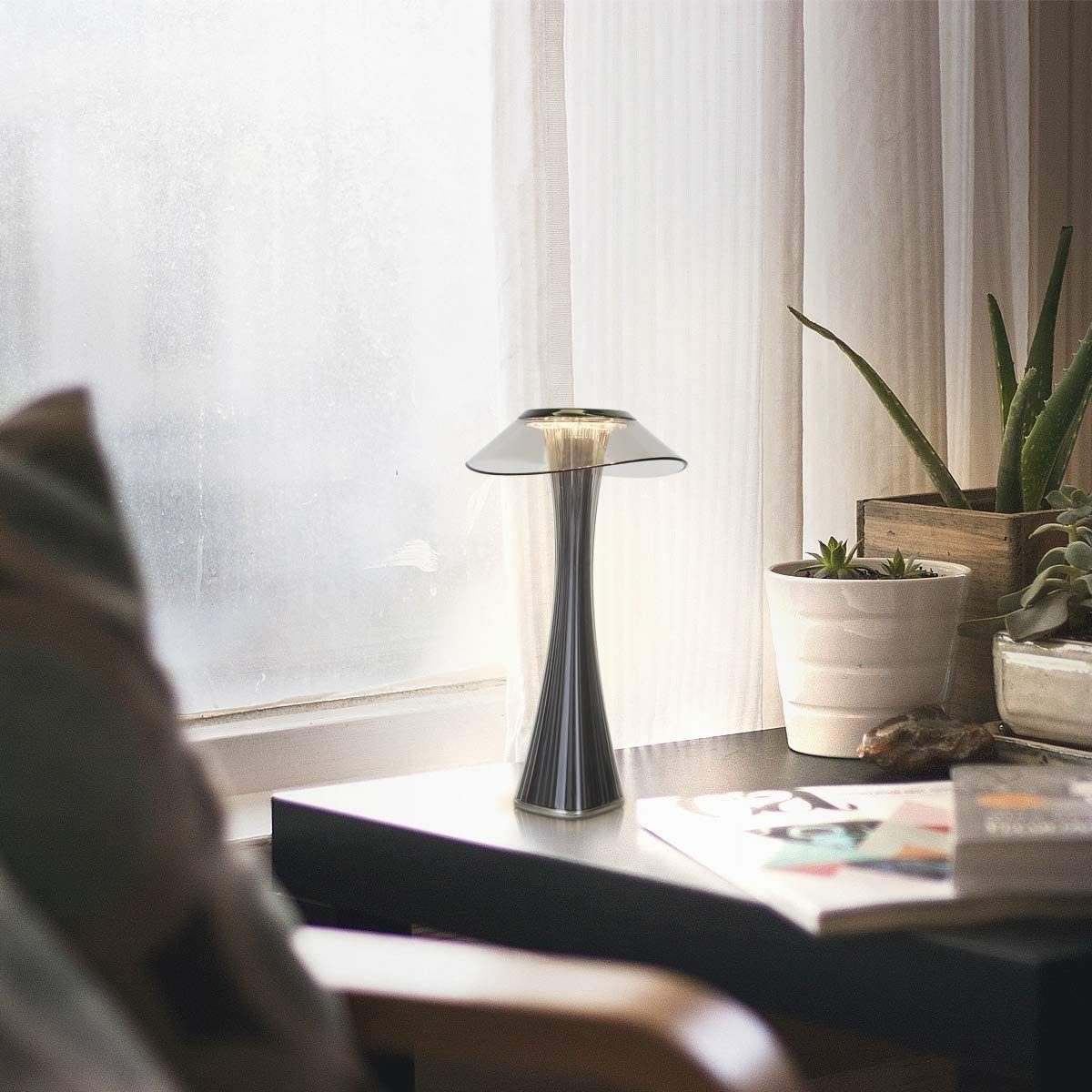 Nowoczesna lampa stołowa z przezroczystą podstawą stoi na biurku w pobliżu okna i rośliny.