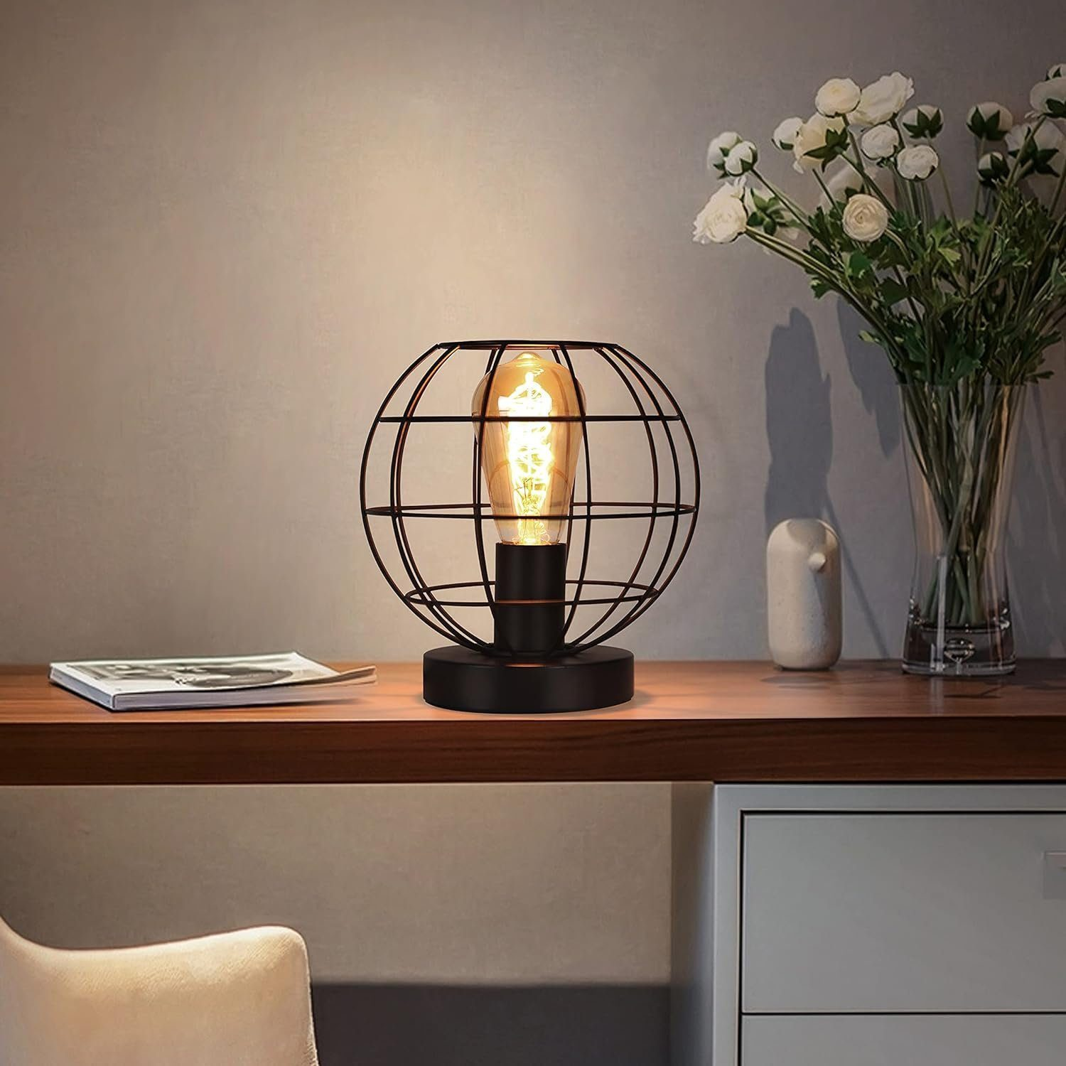 Lampa stołowa z czarnym metalowym kloszem w kształcie klatki i widoczną żarówką. Stoi na biurku obok dekoracji.