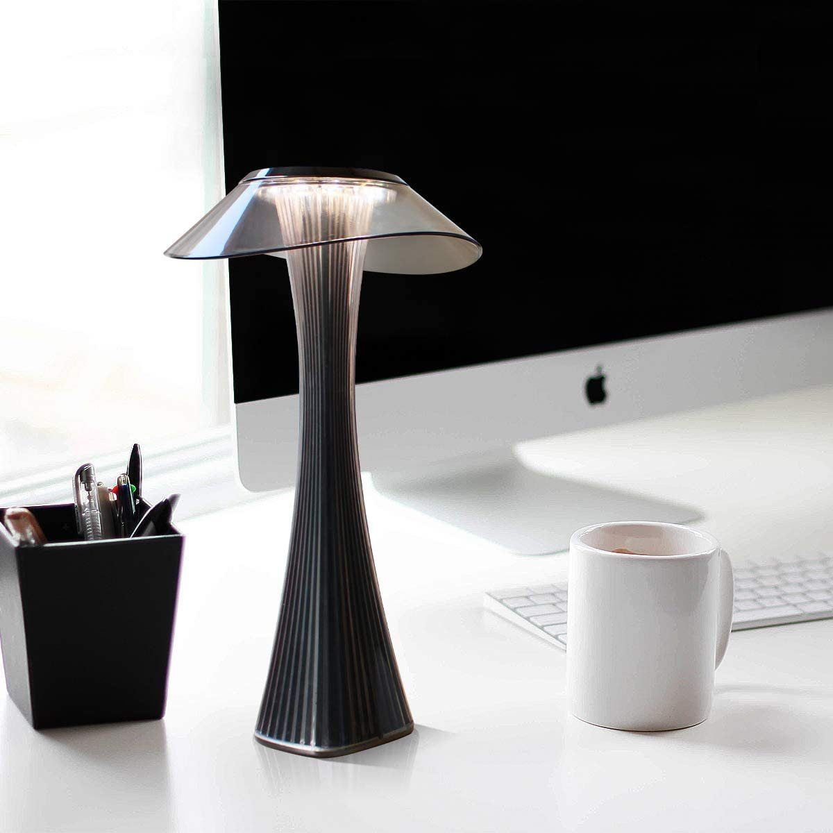 Lampada da tavolo moderna con un design unico. È su una scrivania bianca con penne e una tazza da caffè. Un computer è sullo sfondo.