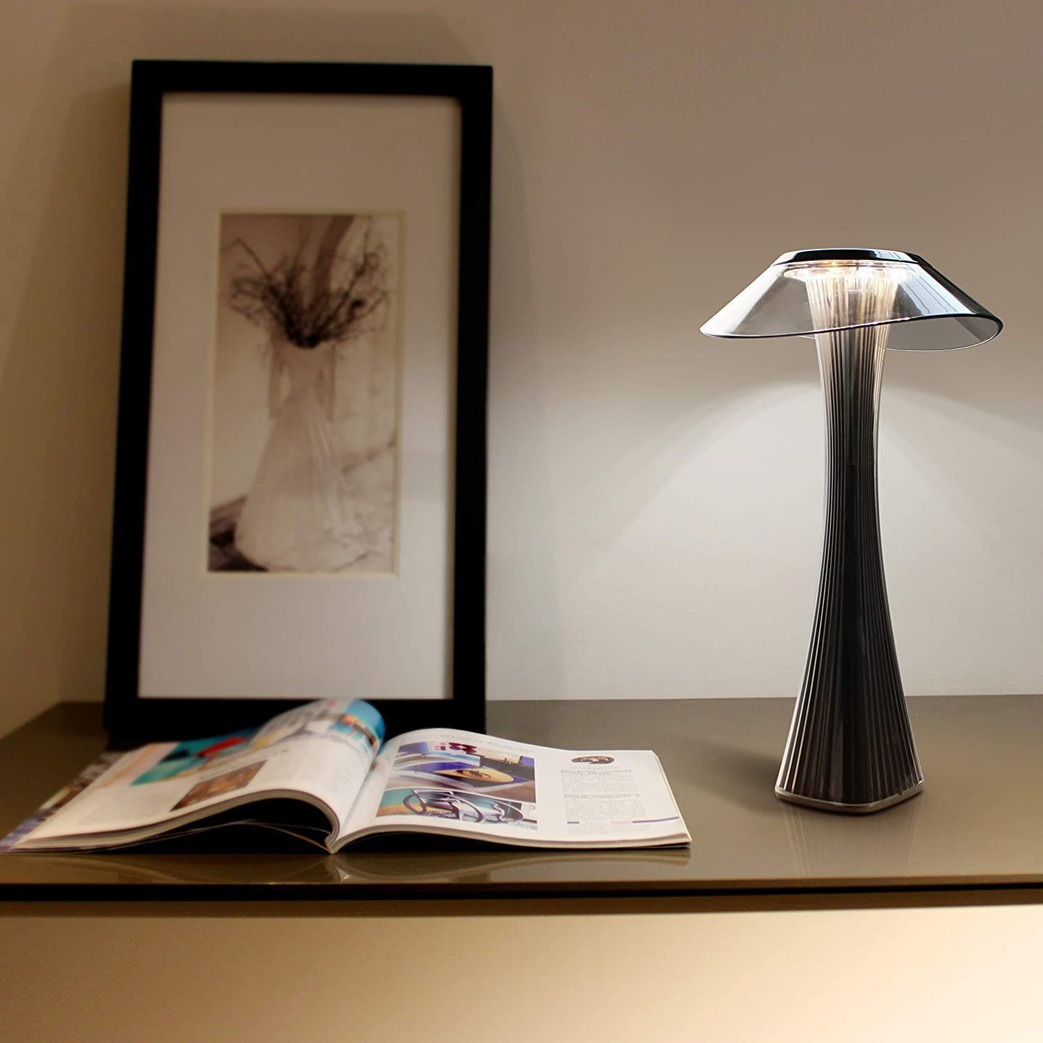Una lampada moderna con una parte superiore trasparente, una base alta e scanalata e un libro aperto su un tavolo vicino a un'immagine incorniciata.