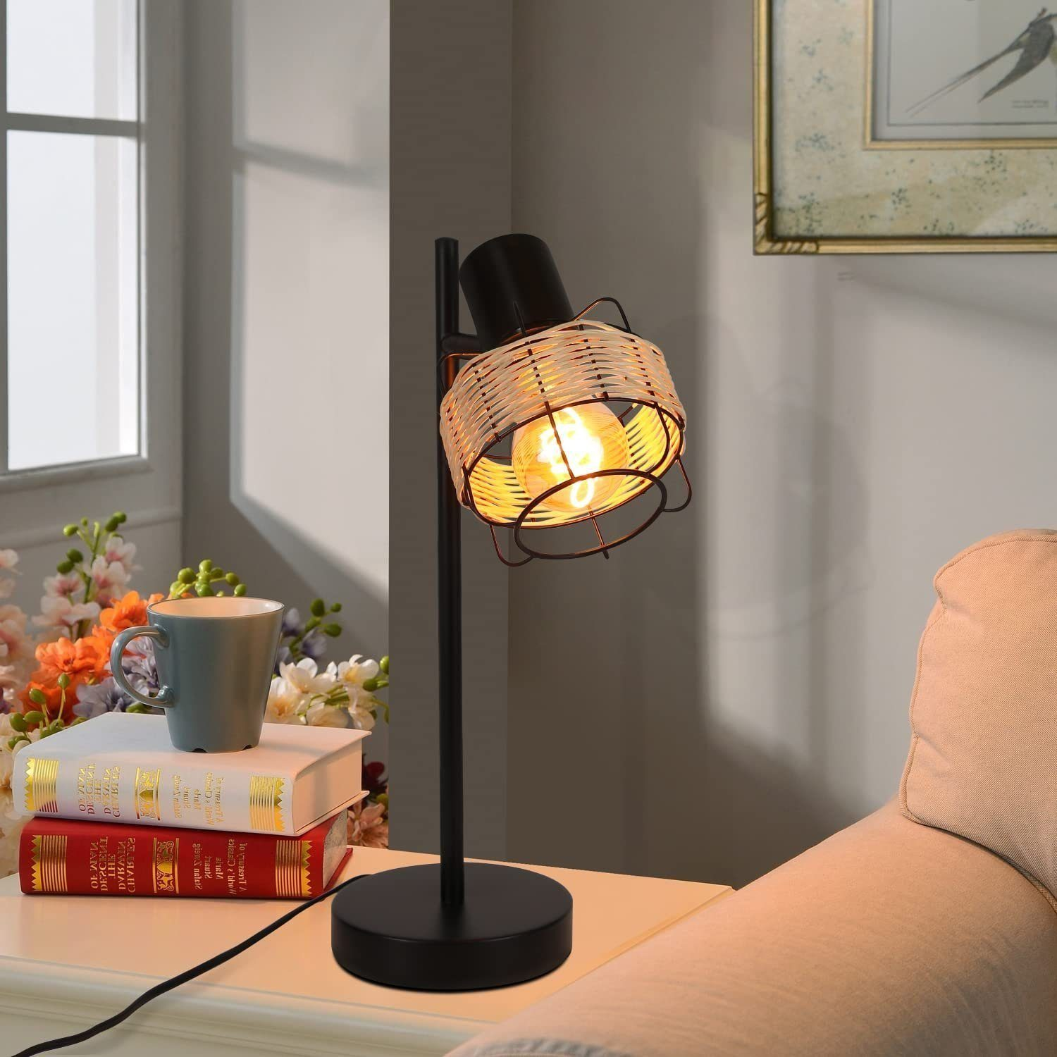 Lampa stołowa z czarną podstawą i wiklinowym kloszem, obok książek i kubka.