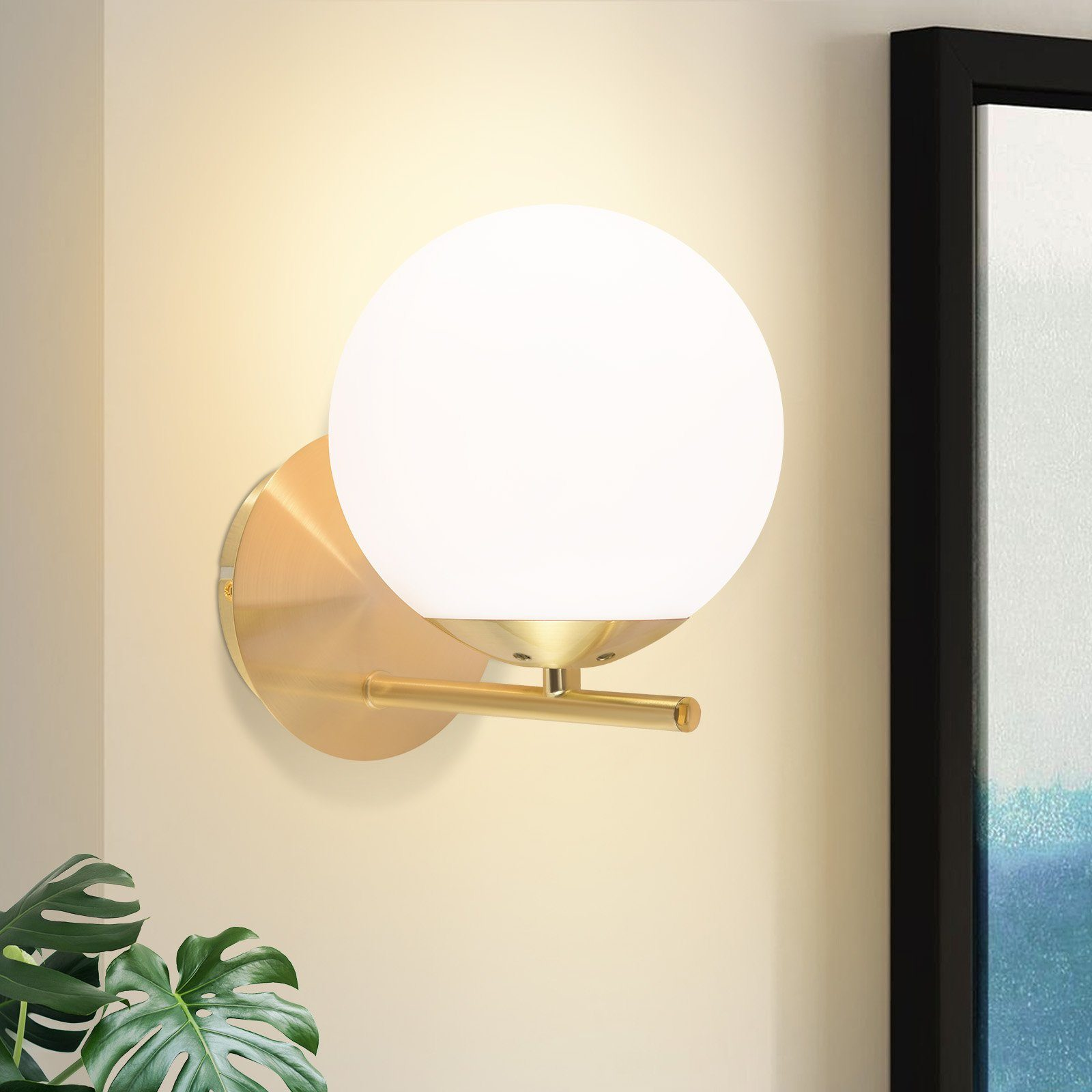 Złoty kinkiet z białą kulą. Jest zamontowany na ścianie. Tło ma czarną ramę.