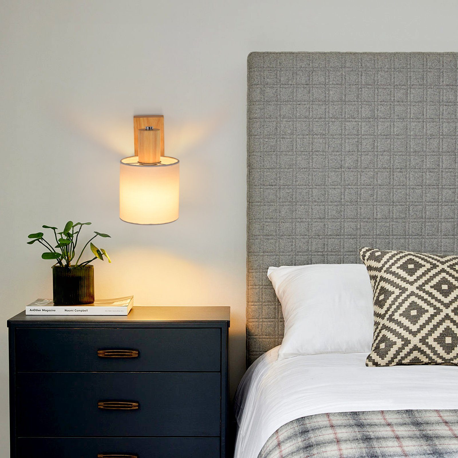 Scena sypialni: lampa na ścianie, stolik nocny z rośliną, łóżko z zagłówkiem i poduszkami.