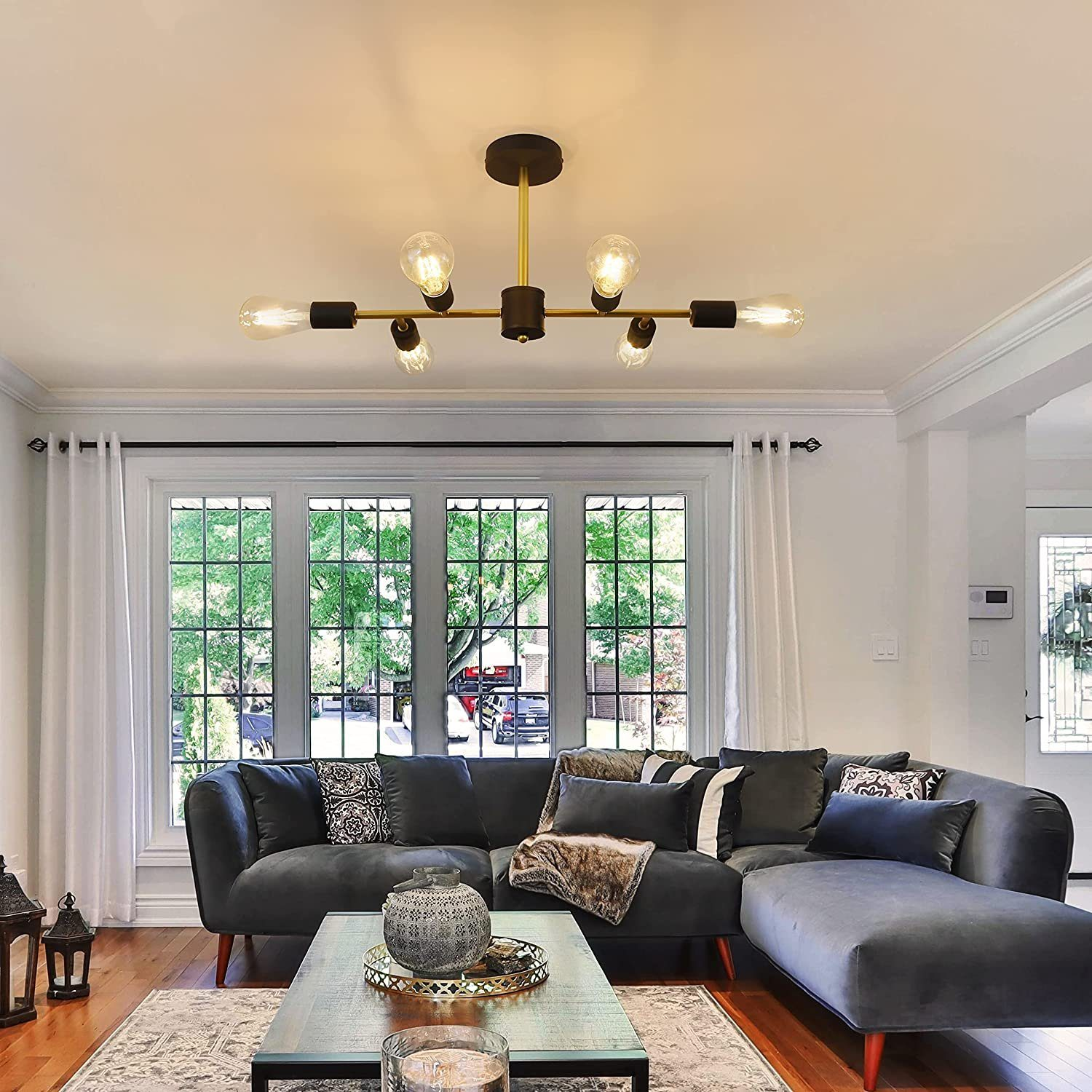 Salon z ciemnoszarą sofą modułową, stolikiem kawowym i żyrandolem z żarówkami Edisona. Duże okna.