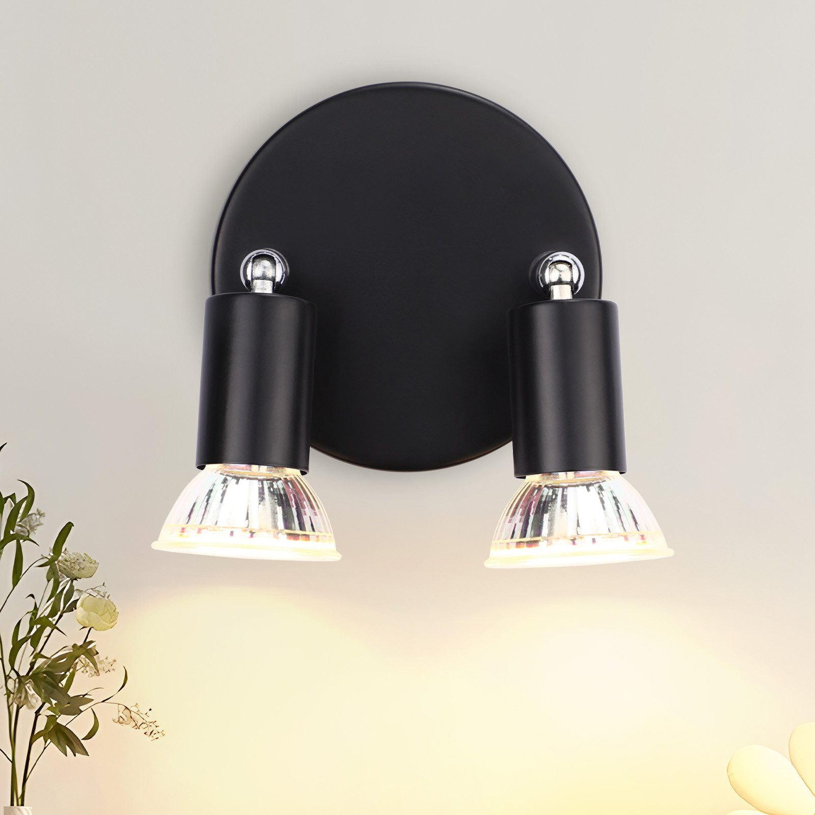 Czarny, dwugłowicowy reflektor na ścianie, oświetlający. Dwie lampy na czarnej, okrągłej podstawie.