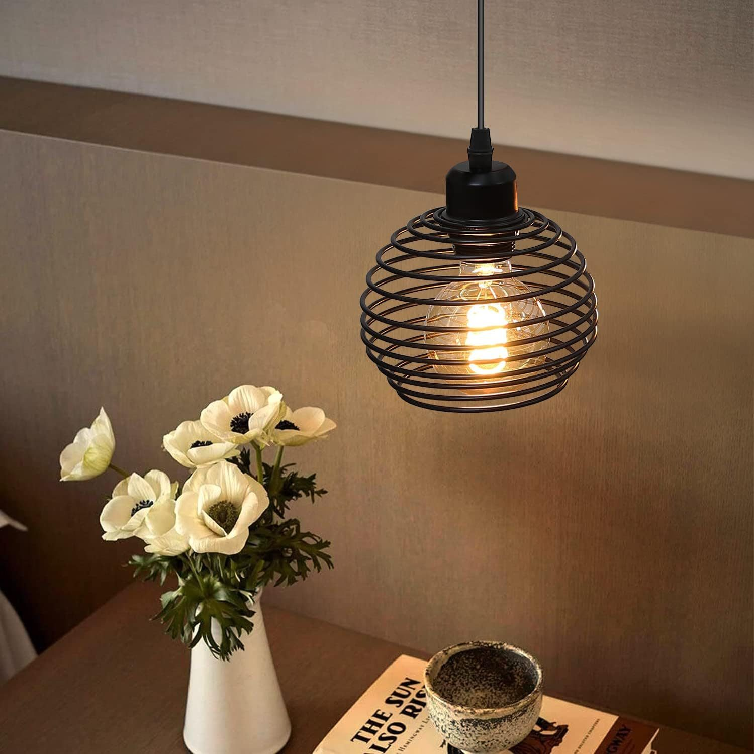 Czarna lampa wisząca z spiralną konstrukcją klatki. Wisi nad stołem z kwiatami i książką.