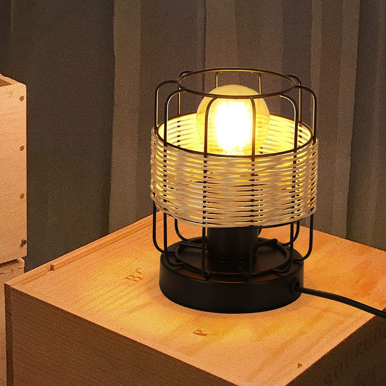 Lampa stołowa na drewnianej powierzchni. Lampa ma czarną podstawę, tkany klosz i świecącą żarówkę.