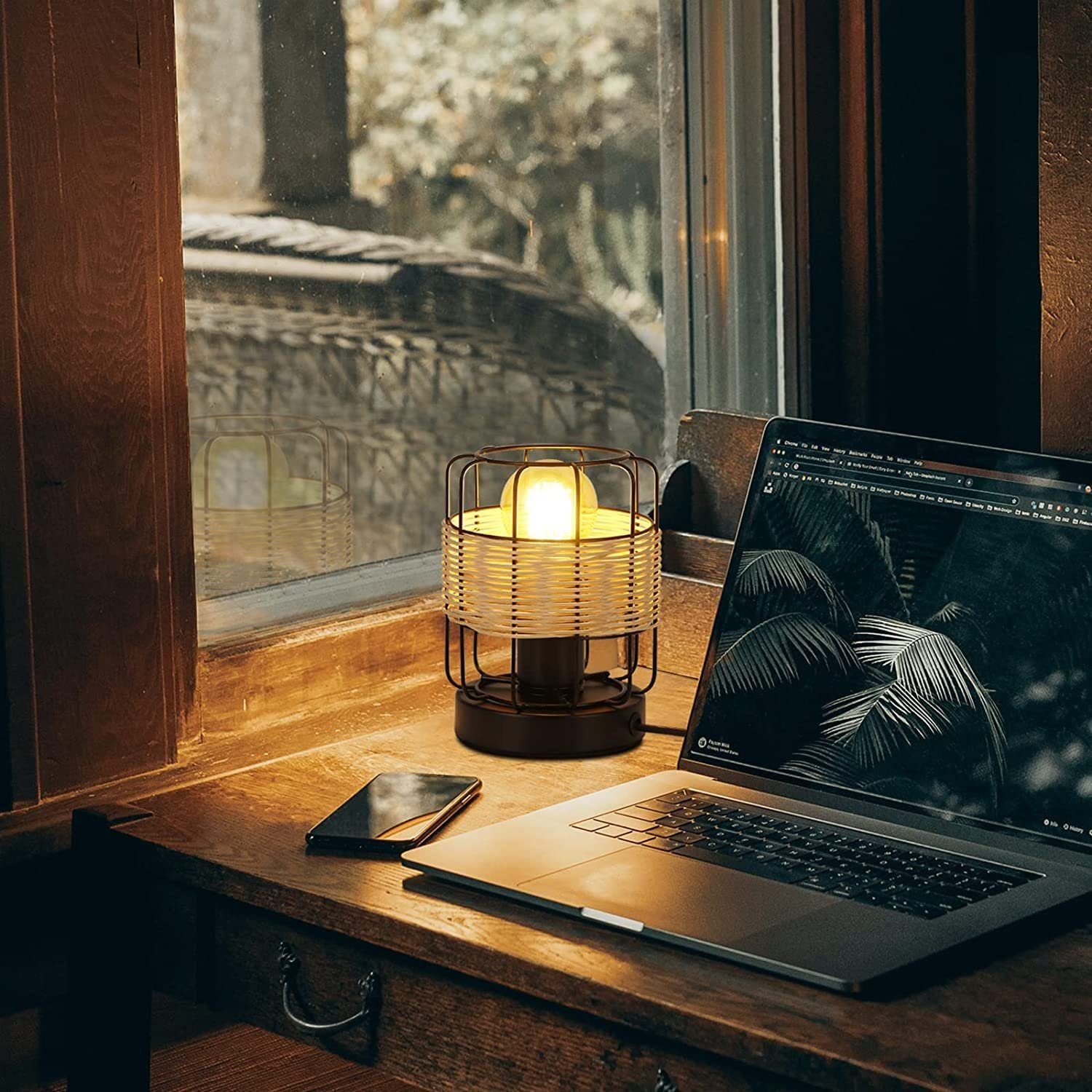 Zapalona lampa stoi na drewnianym biurku obok laptopa i telefonu. W tle okno.