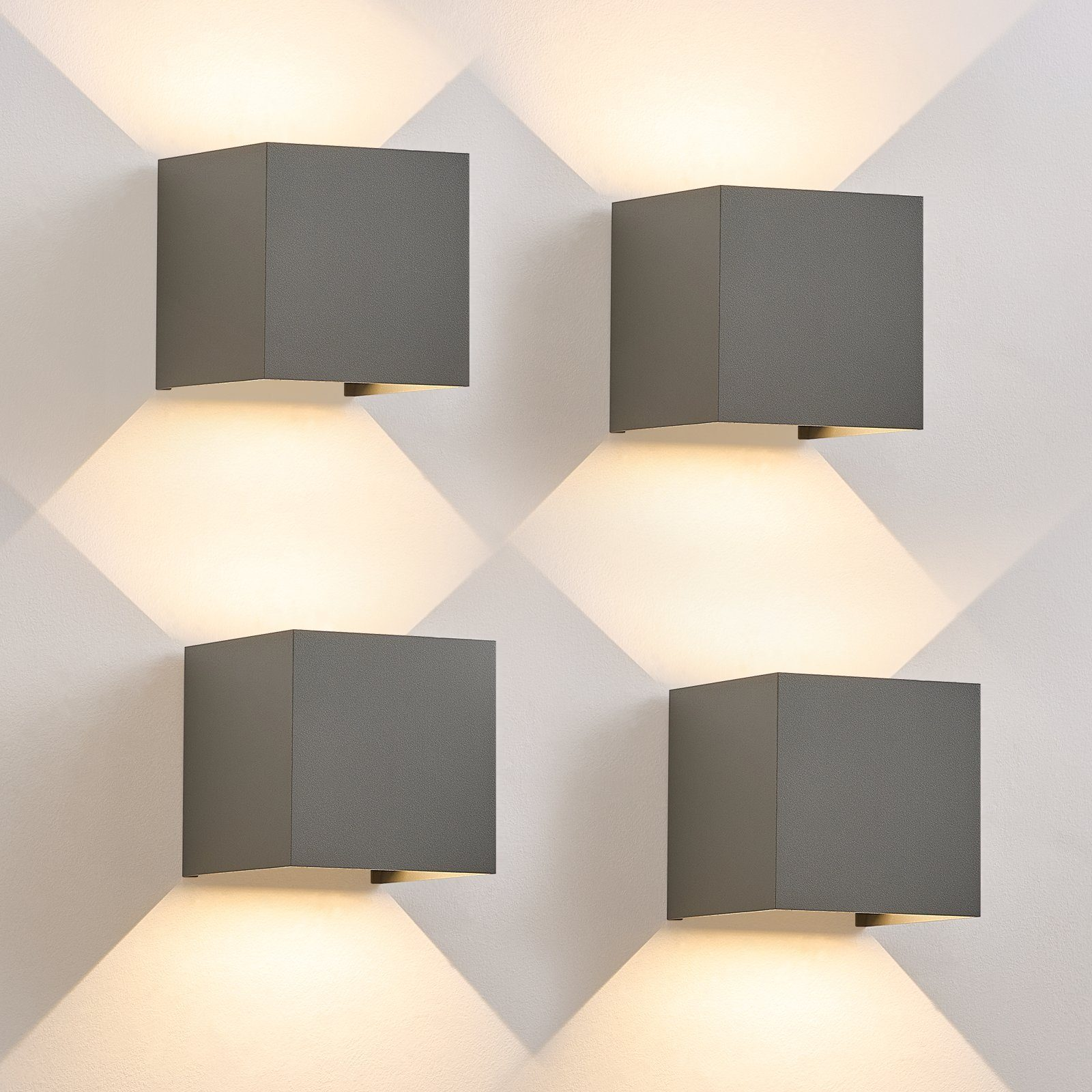 Cztery szare lampy ścienne w kształcie kostki. Każda emituje światło w dół, oświetlając ścianę.