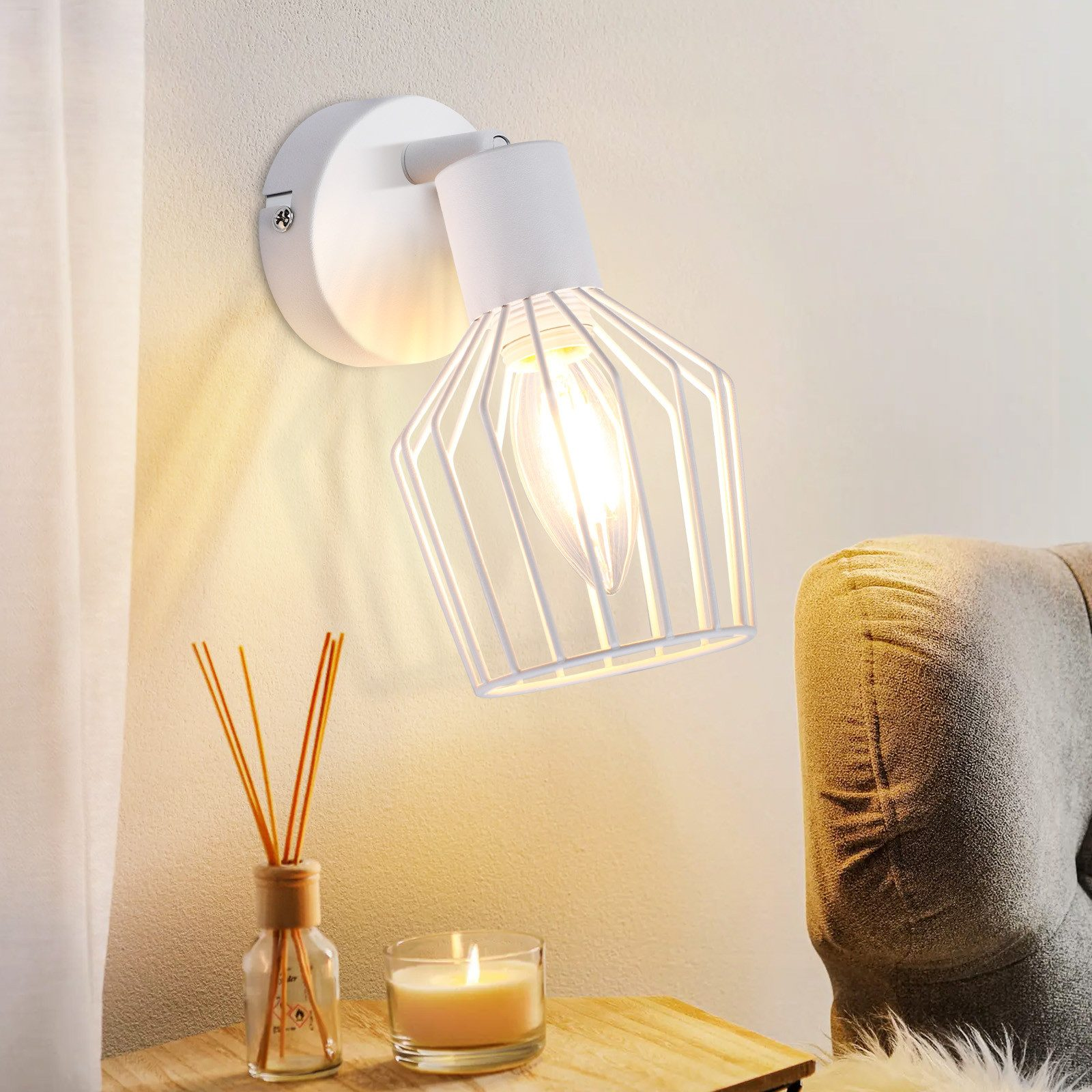 Una lampada da parete bianca con paralume a gabbia metallica, che proietta la luce su un tavolo di legno con una candela.