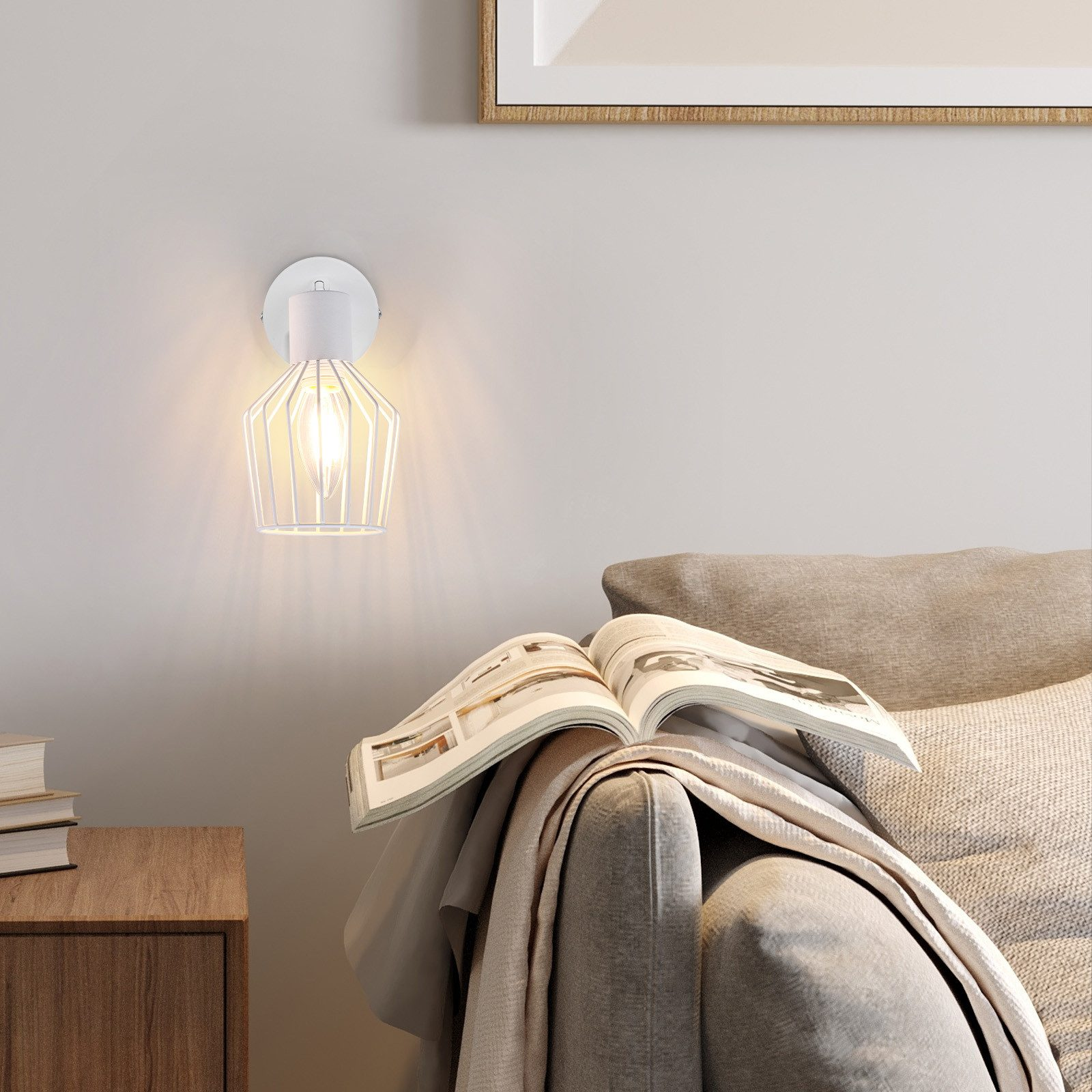 Biała lampa ścienna z kloszem w kształcie klatki, zapalona, obok sofy z książką i kocem.