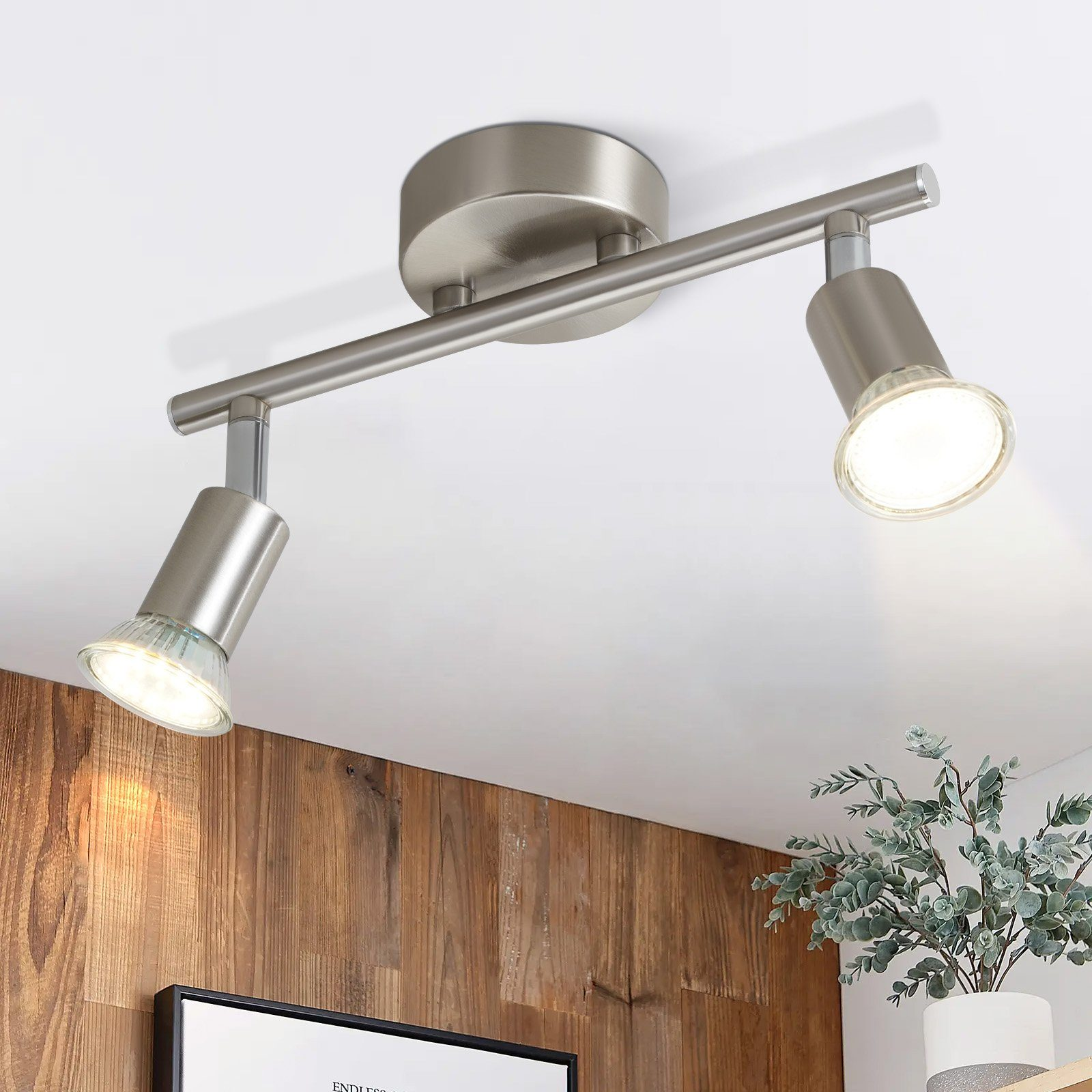 Srebrna lampa sufitowa z dwoma regulowanymi reflektorami, oświetlająca drewnianą ścianę. Roślina znajduje się na pierwszym planie.