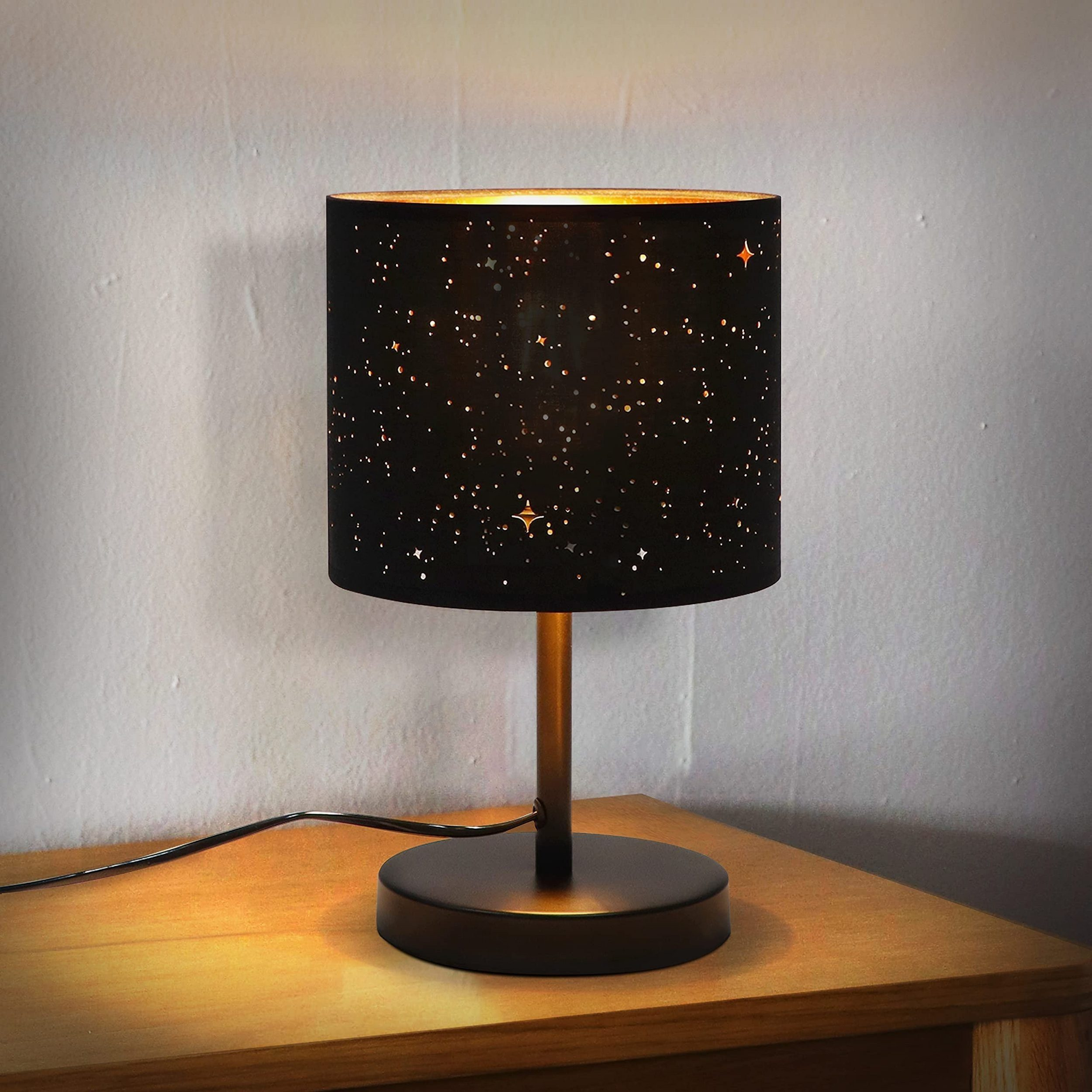 Lampa z gwieździstą nocą na drewnianym stole. To cylindryczna lampa z ciemnym tłem i białymi wzorami gwiazd.