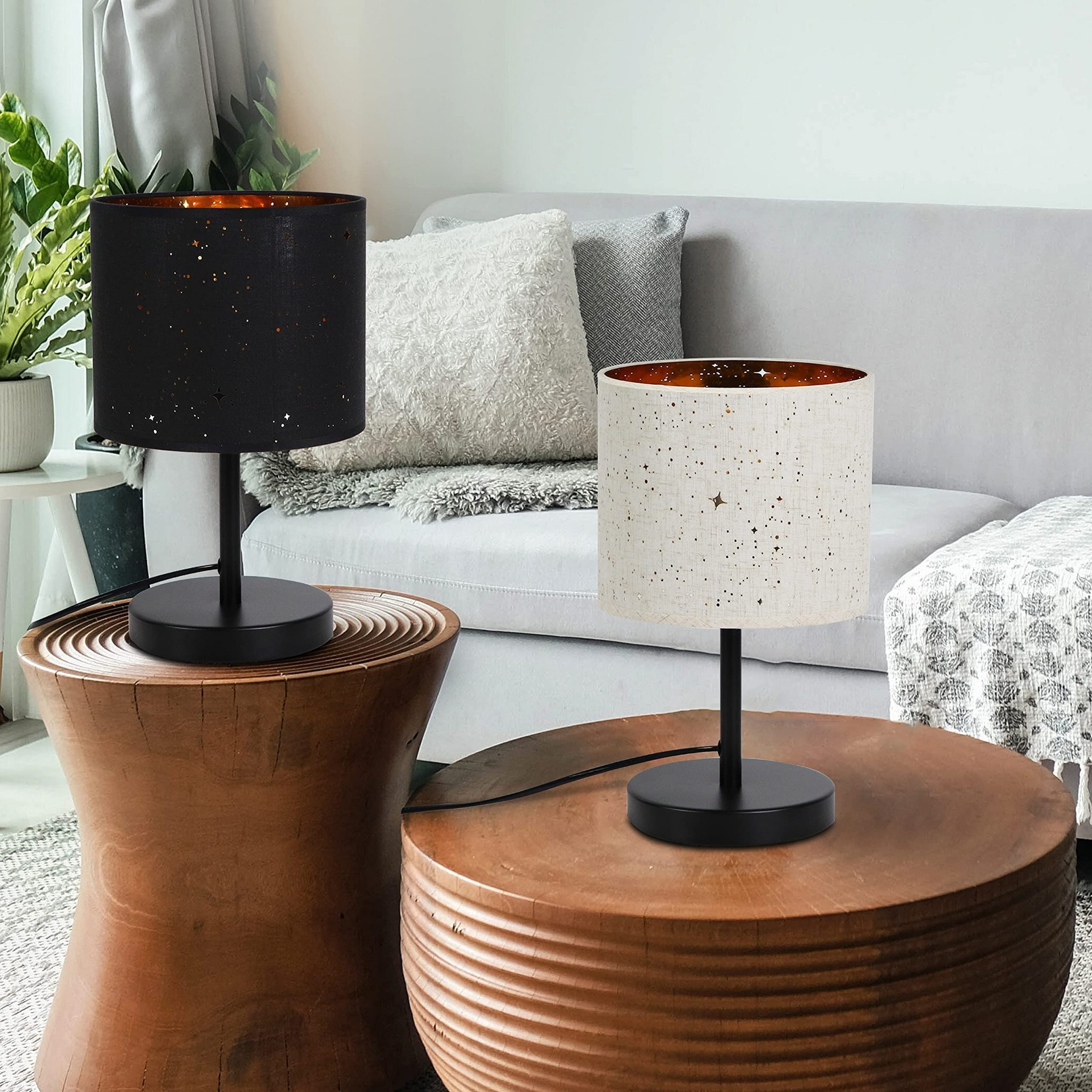 Dwie lampy stołowe, jedna czarna ze wzorem gwiazd, druga biała ze wzorem gwiazd, na drewnianych stolikach obok szarej sofy.