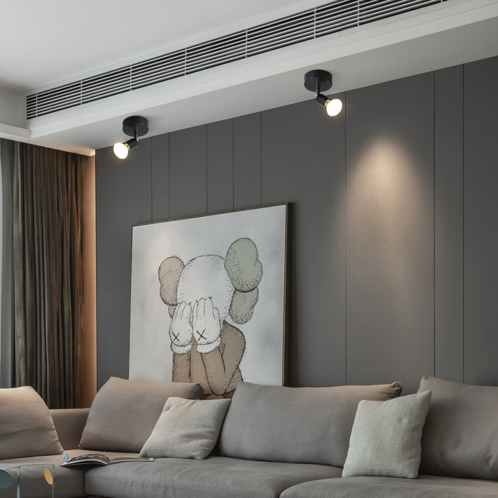 Dwa czarne reflektory zamontowane na białym suficie, oświetlające salon z szarą sofą i sztuką.