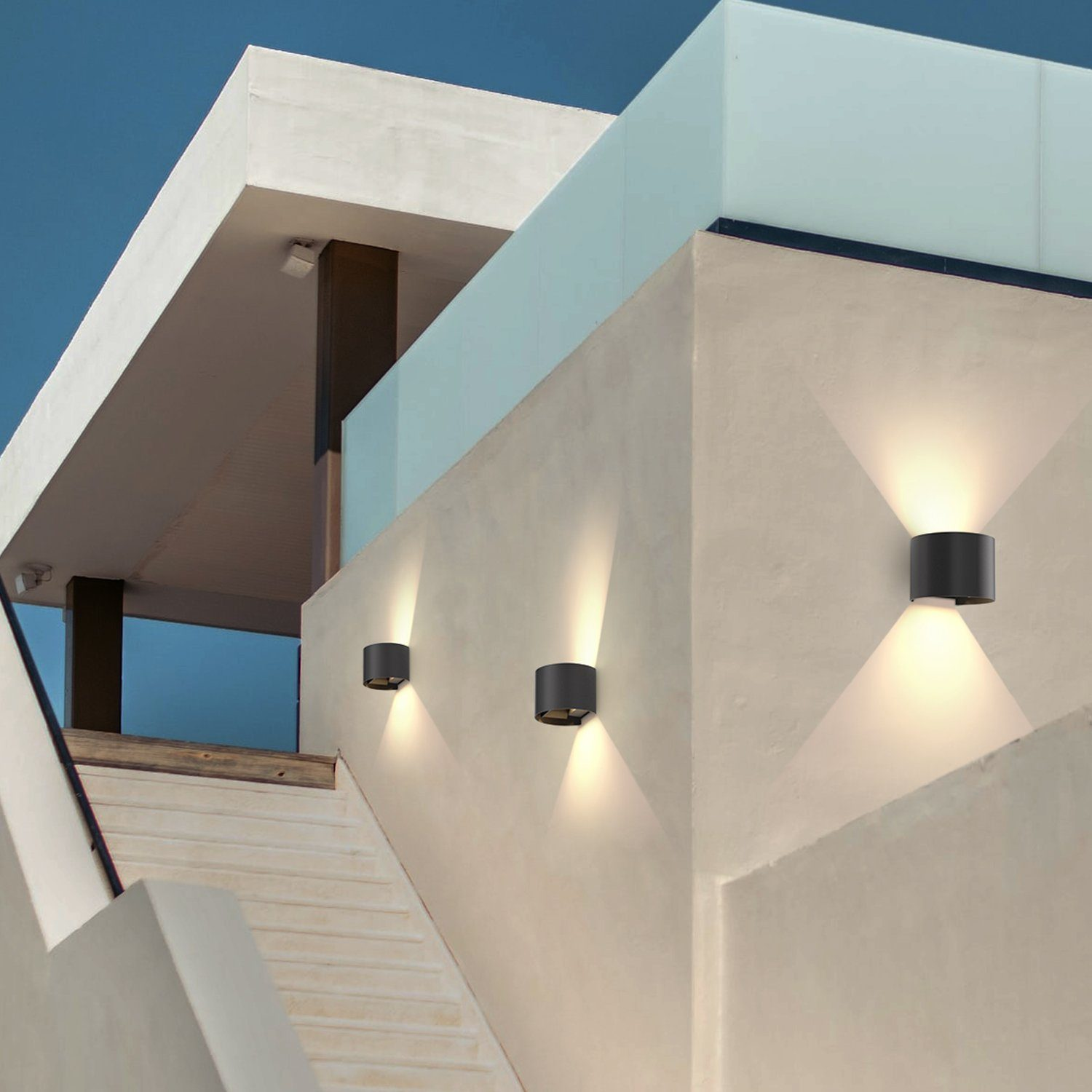Tre luci da parete nere per esterni su un muro bianco. Illuminano verso il basso.