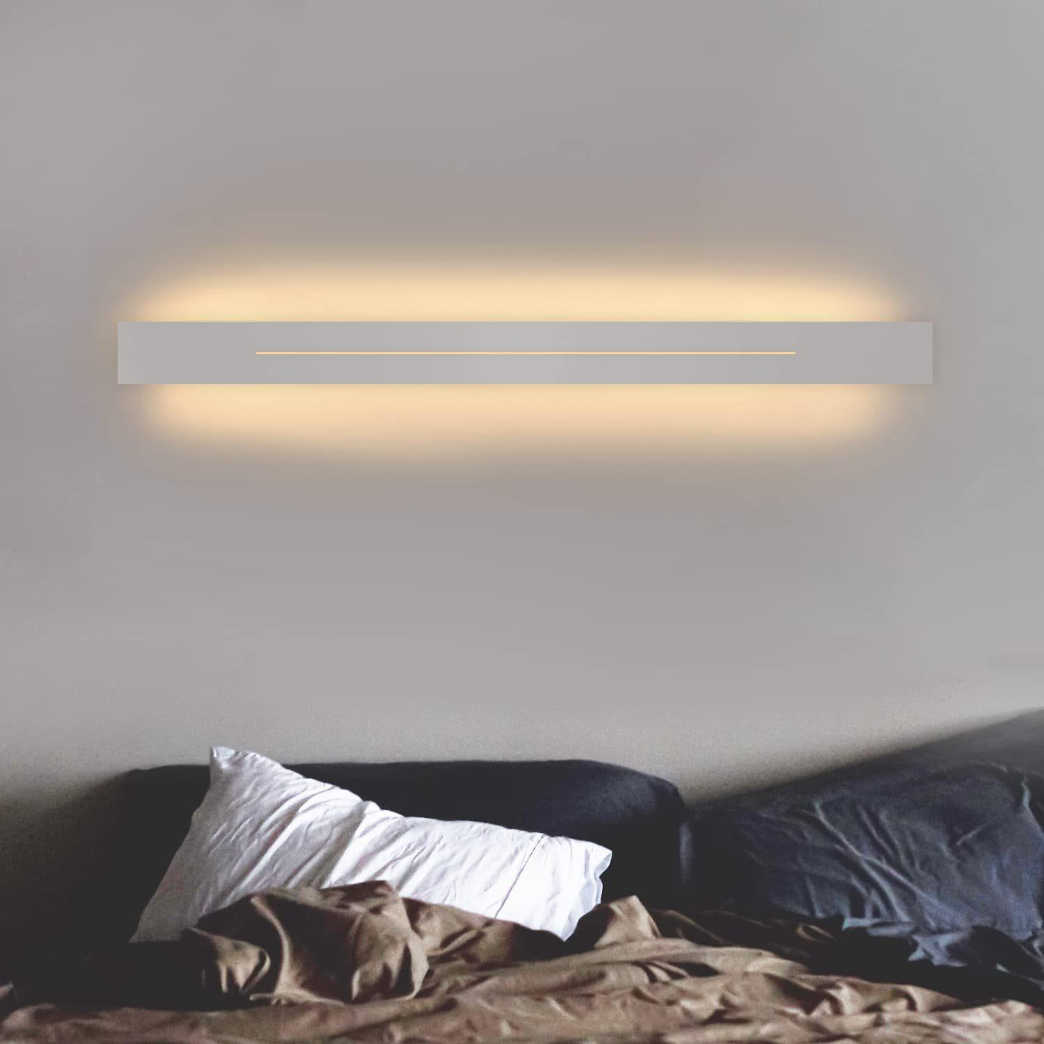 Lampada da parete bianca su muro grigio. Letto con cuscini e coperta visibile sotto.