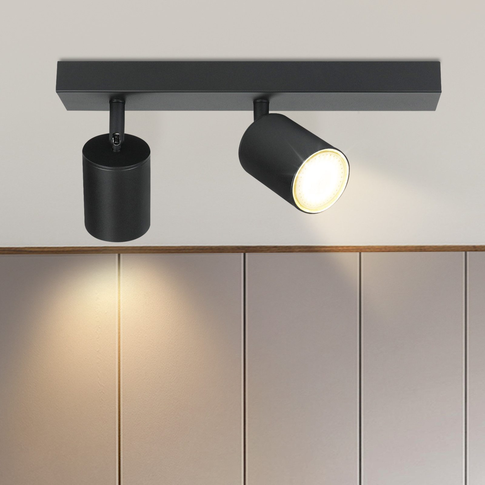 Una lampada da soffitto con due faretti neri. Lo sfondo mostra un muro di colore neutro.