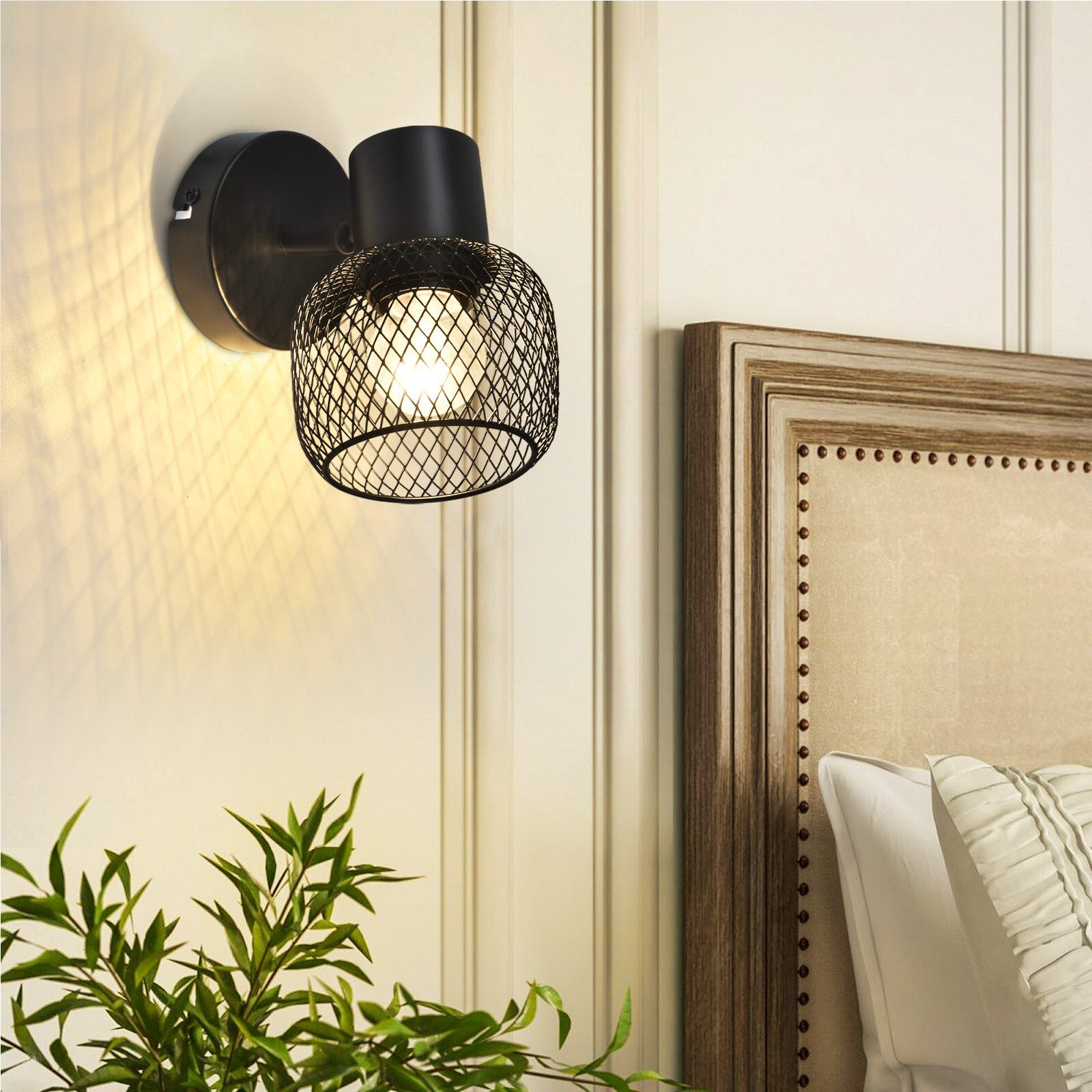 Lampada da parete in metallo nero con paralume a gabbia. Una luce brilla sul muro. Piante verdi in primo piano.