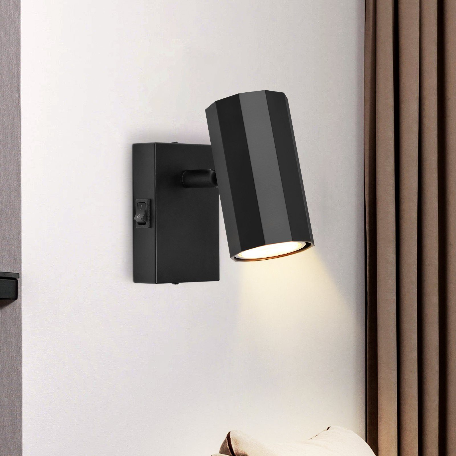 Una lampada da parete nera con interruttore e fascio di luce. È montata su una parete bianca.