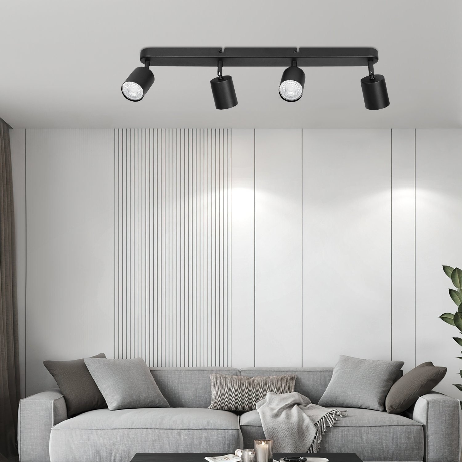 Faretti neri a soffitto illuminano un divano grigio. Sfondo parete bianca. Una pianta sullo sfondo.