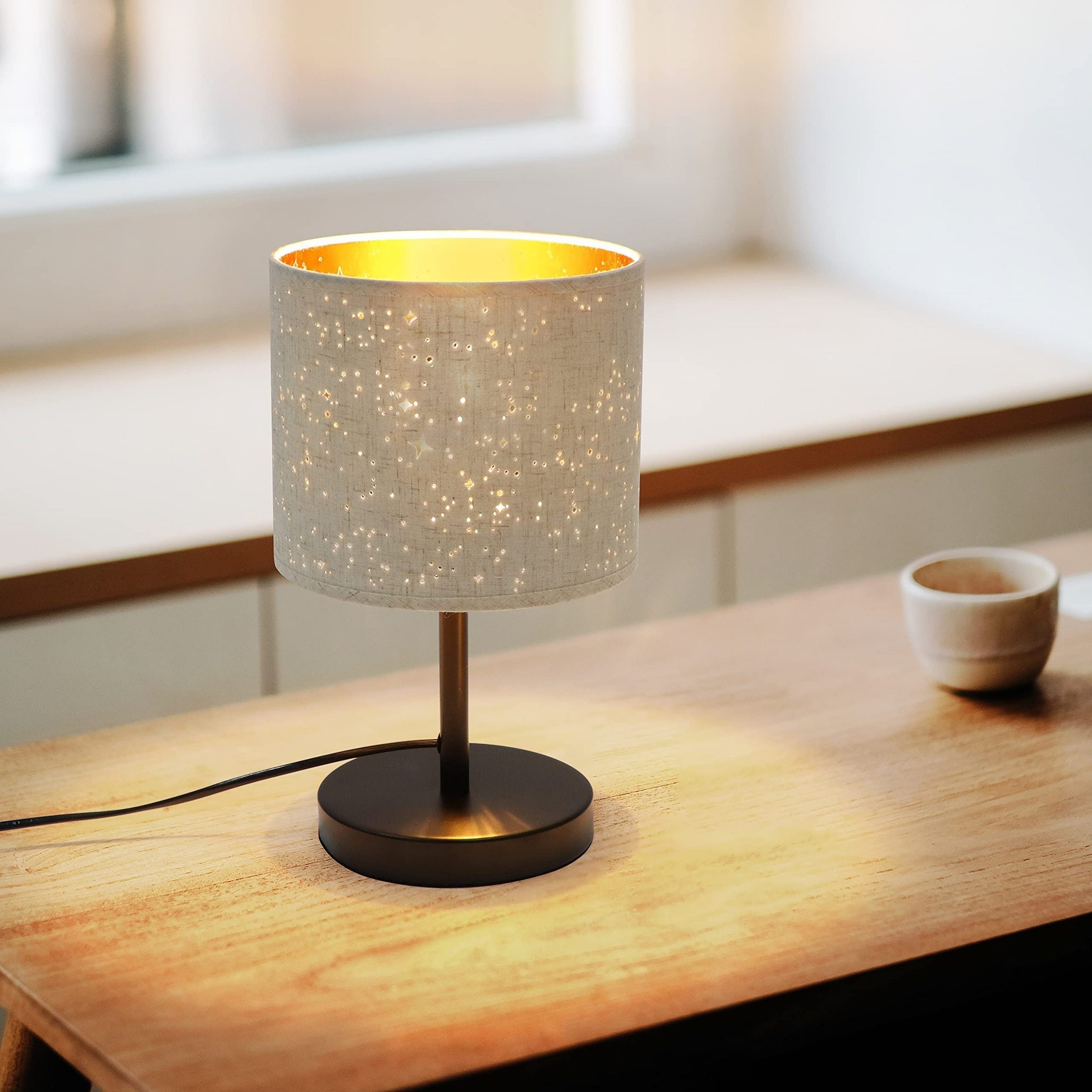 Lampa na drewnianym stole. Ma biały, kropkowany klosz i brązowy stojak. Obok stoi filiżanka.