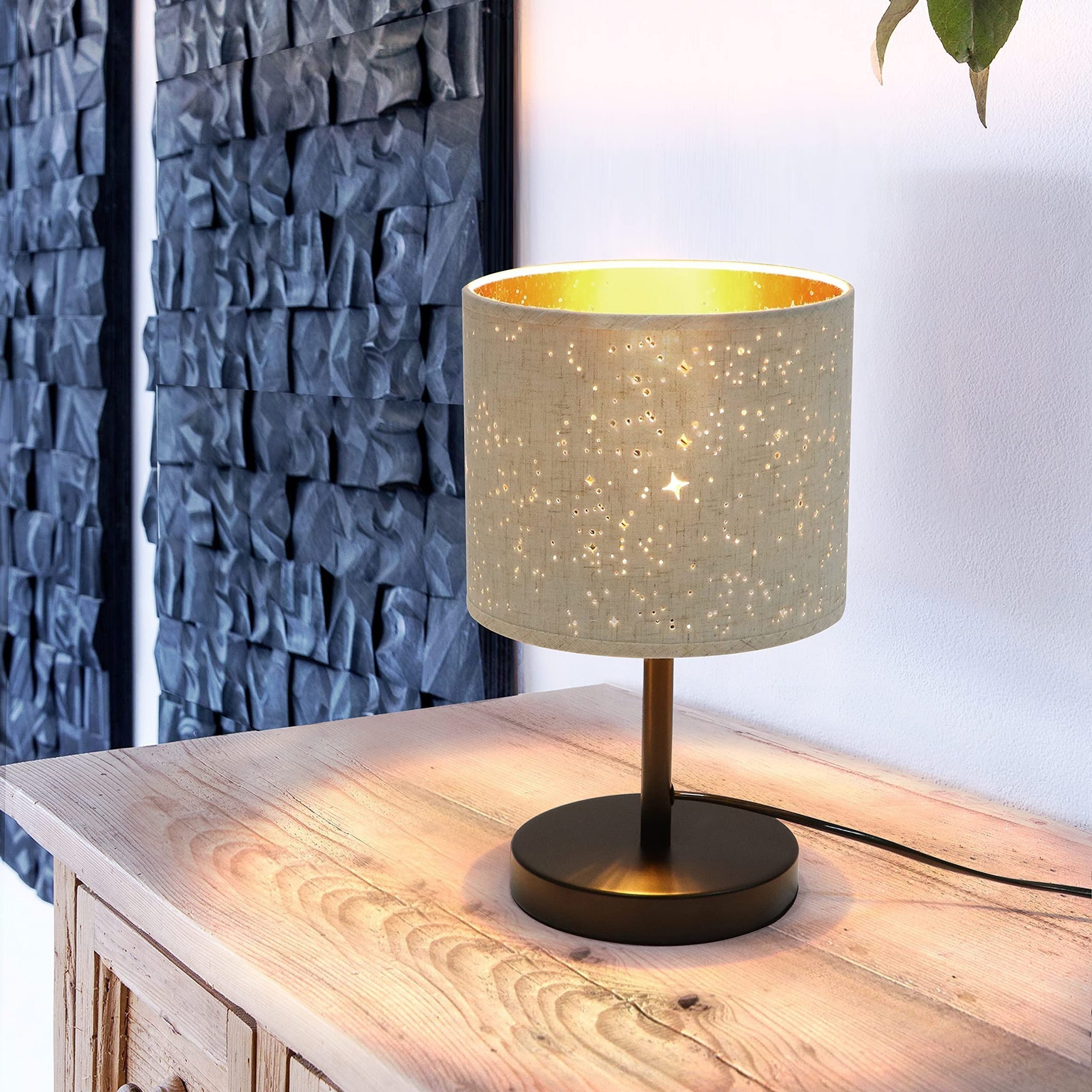 Lampa stołowa z beżowym kloszem w gwiazdki i złotym wnętrzem. Stoi na drewnianym stole.