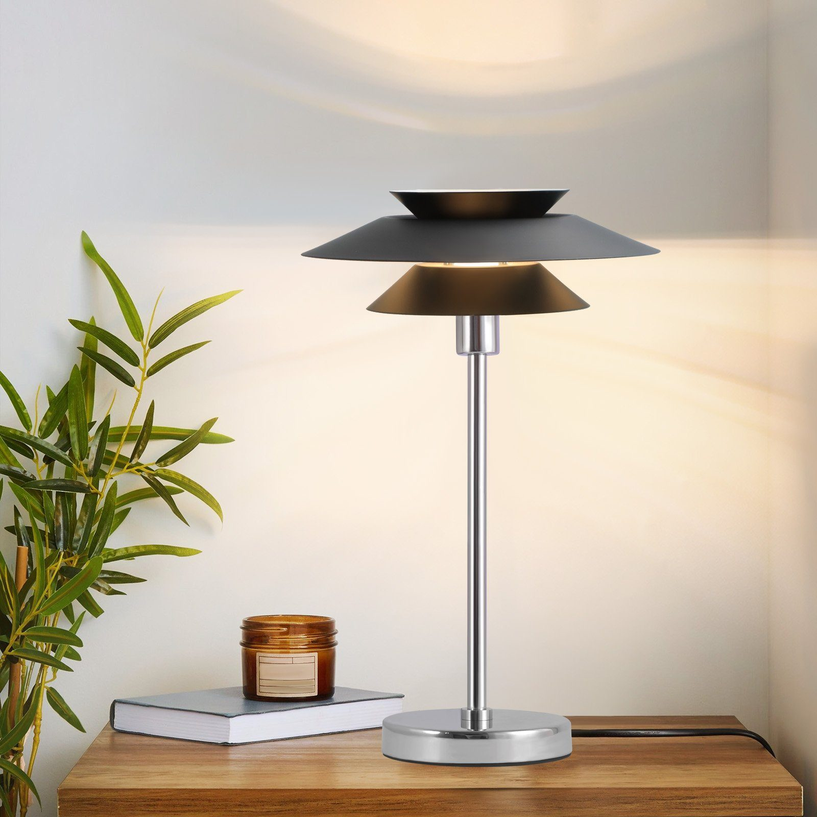 Lampa stołowa z warstwowym, czarnym kloszem i srebrną podstawą, na drewnianym stole obok rośliny i świecy.