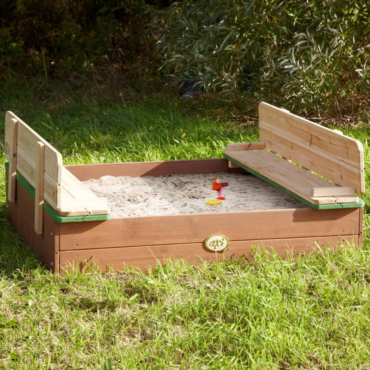 Ein hölzerner Sandkasten mit Sand, zwei Bänken und einem kleinen roten und gelben Spielzeug.