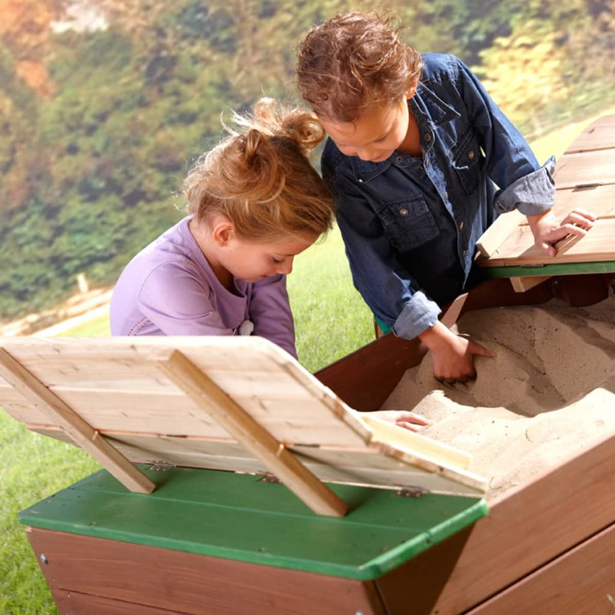 Zwei Kinder spielen in einem Sandkasten. Ein Kind trägt ein Jeanshemd. Der Sandkasten ist aus Holz.