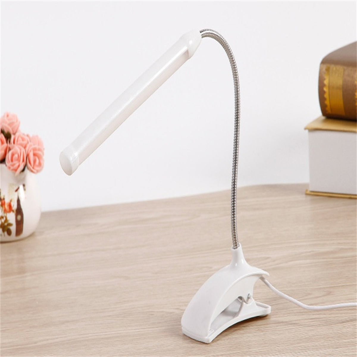 Eine weiße Schreibtischlampe mit flexiblem Hals. Sie ist an einem Holztisch befestigt. Auf dem Tisch liegen Bücher.