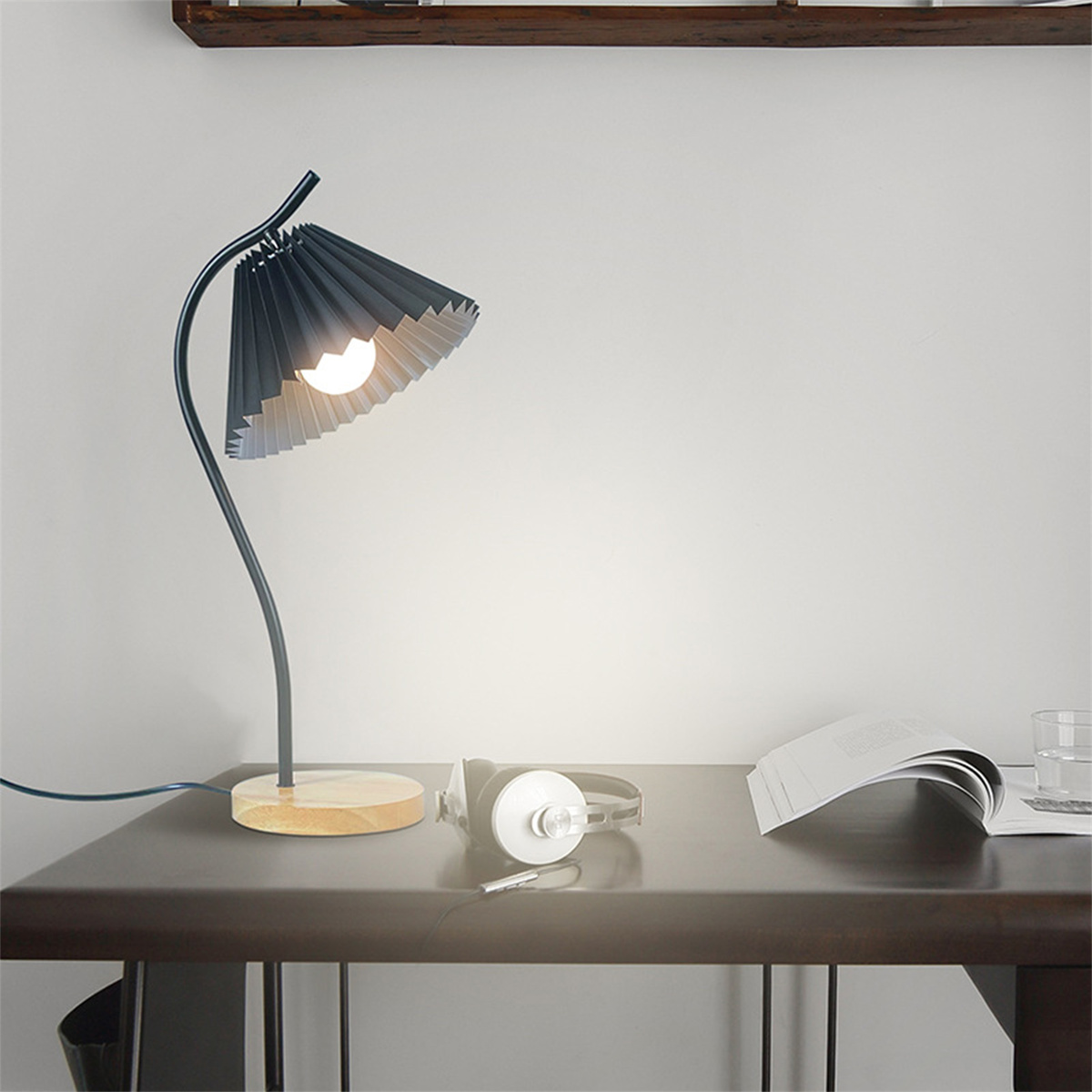 Eine moderne Schreibtischlampe mit schwarzem Schirm und Holzfuß steht auf einem dunklen Schreibtisch. Kopfhörer und ein Buch liegen auf dem Tisch.