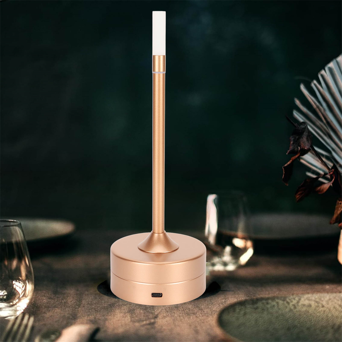 Goldfarbene Lampe mit hohem, schlankem Körper und Sockel. Sie steht auf einem Holztisch mit Gläsern.