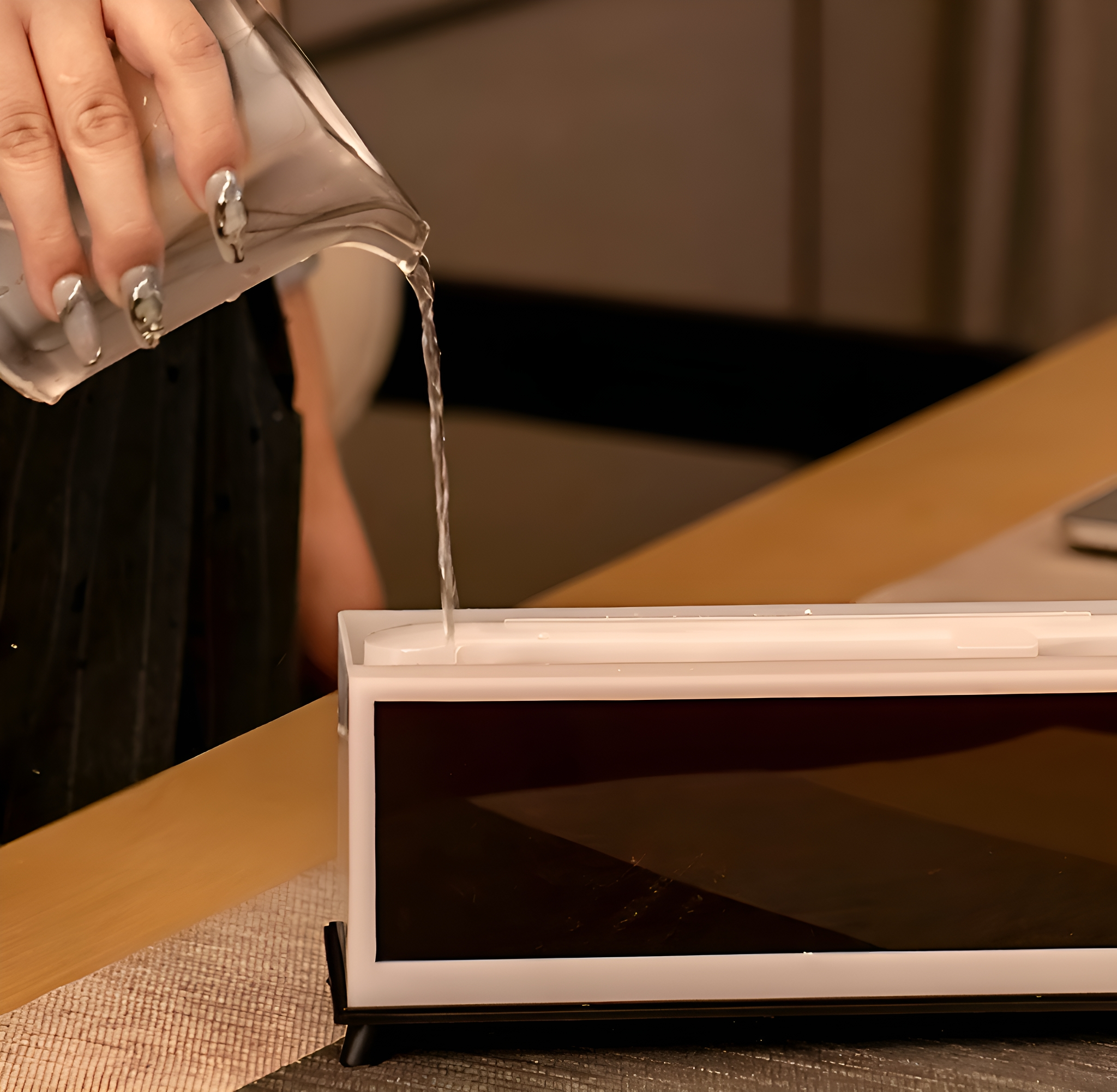 Eine Person gießt Wasser aus einem Glas in einen weißen und schwarzen rechteckigen Behälter. Der Behälter steht auf einer Holzoberfläche.