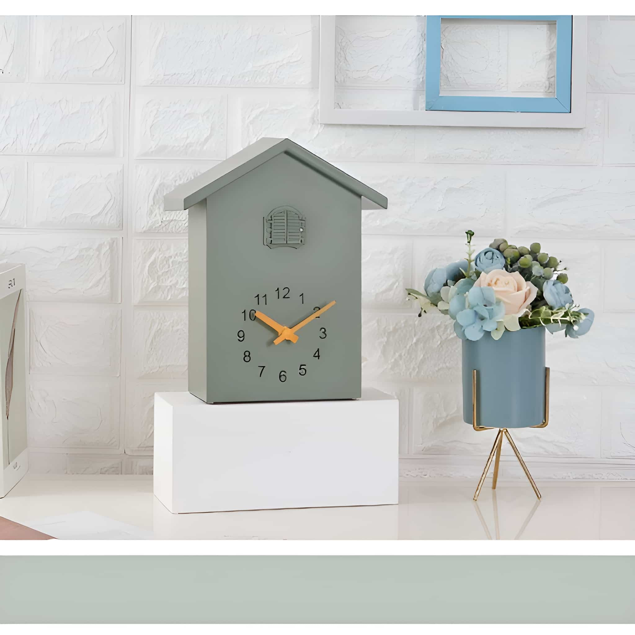 Eine graue Uhr in Form eines Hauses steht auf einer weißen Oberfläche. Blumen in einer blauen Vase sind in der Nähe.