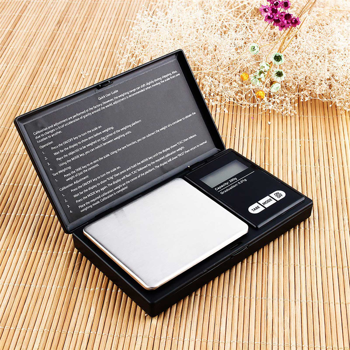 Geöffnete schwarze Digitalwaage mit Anweisungen und kleinem Blumengesteck im Hintergrund.