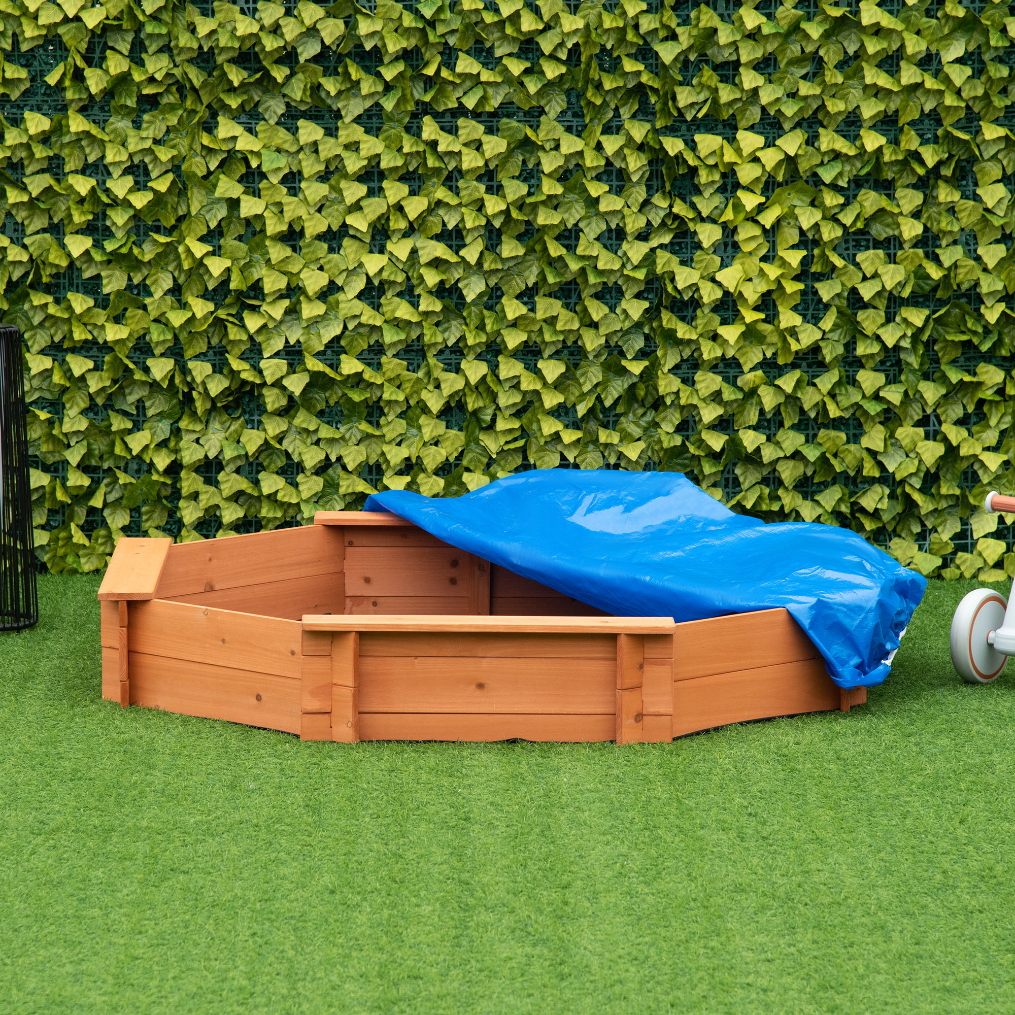 Una sabbiera in legno con telone blu su erba verde, con sfondo di foglie.