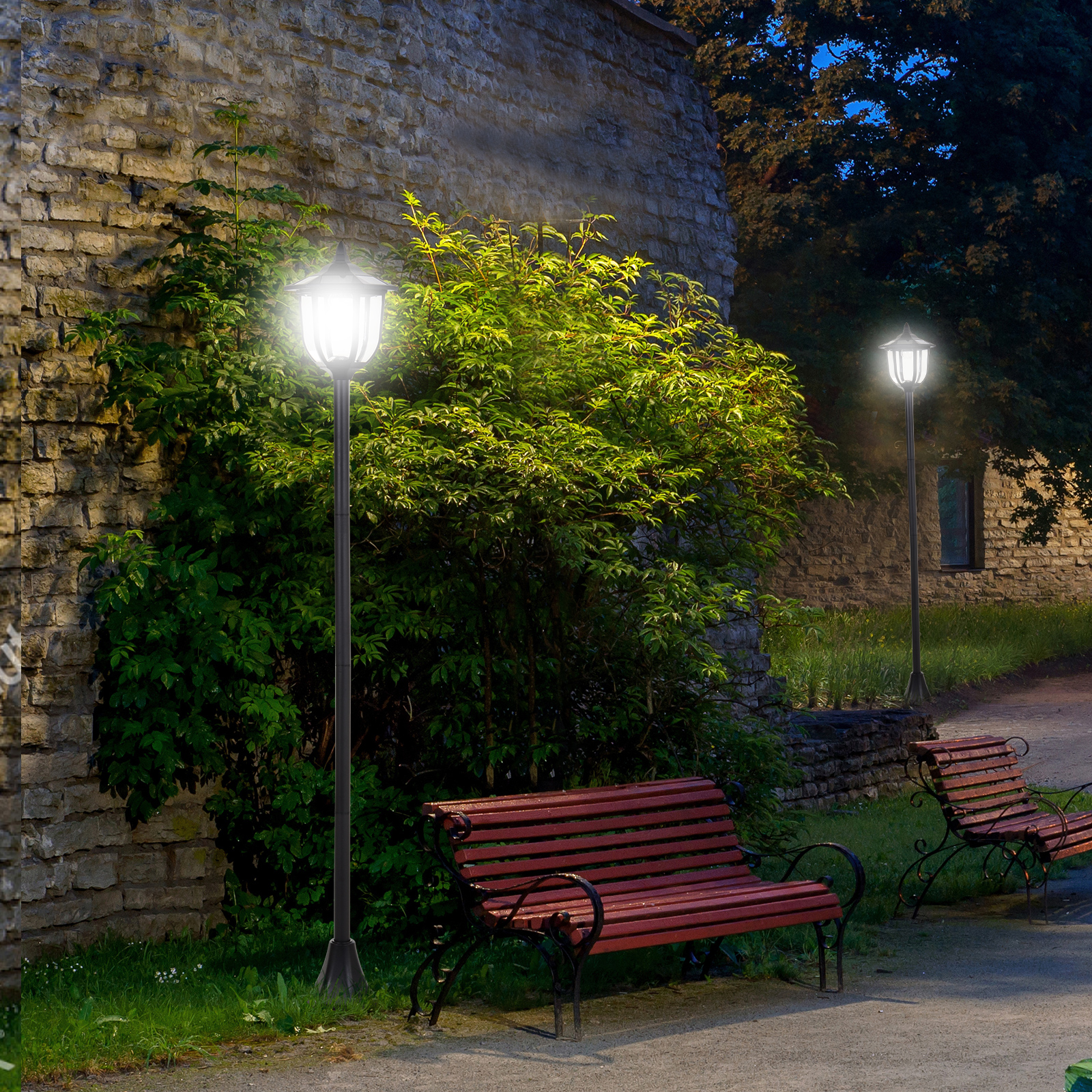 Due lampioni illuminano un muro di pietra e panchine. Arbusti verdi circondano le lampade.