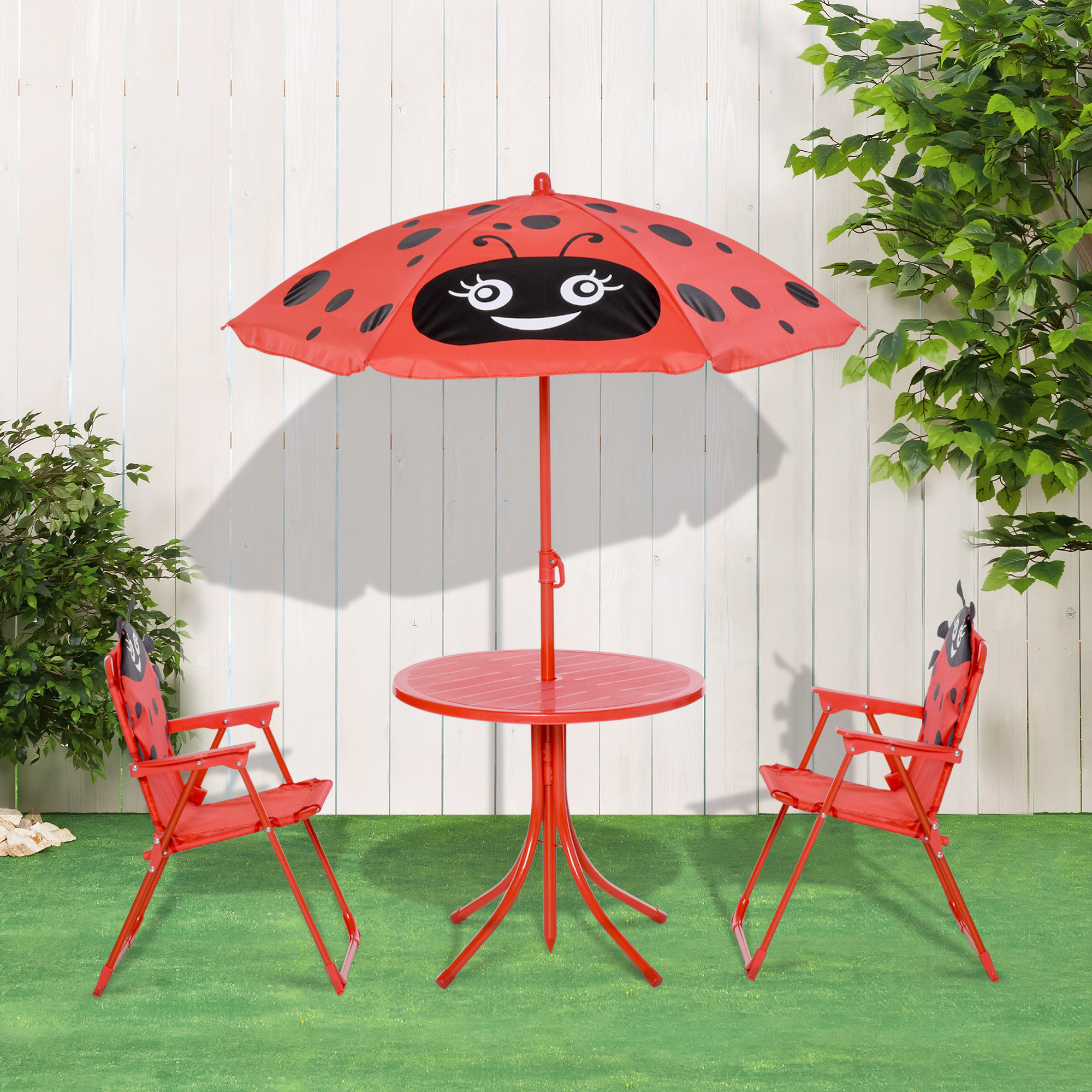Set tavolo da esterno rosso per bambini con ombrellone a coccinella, due sedie sull'erba, sfondo recinzione bianca.