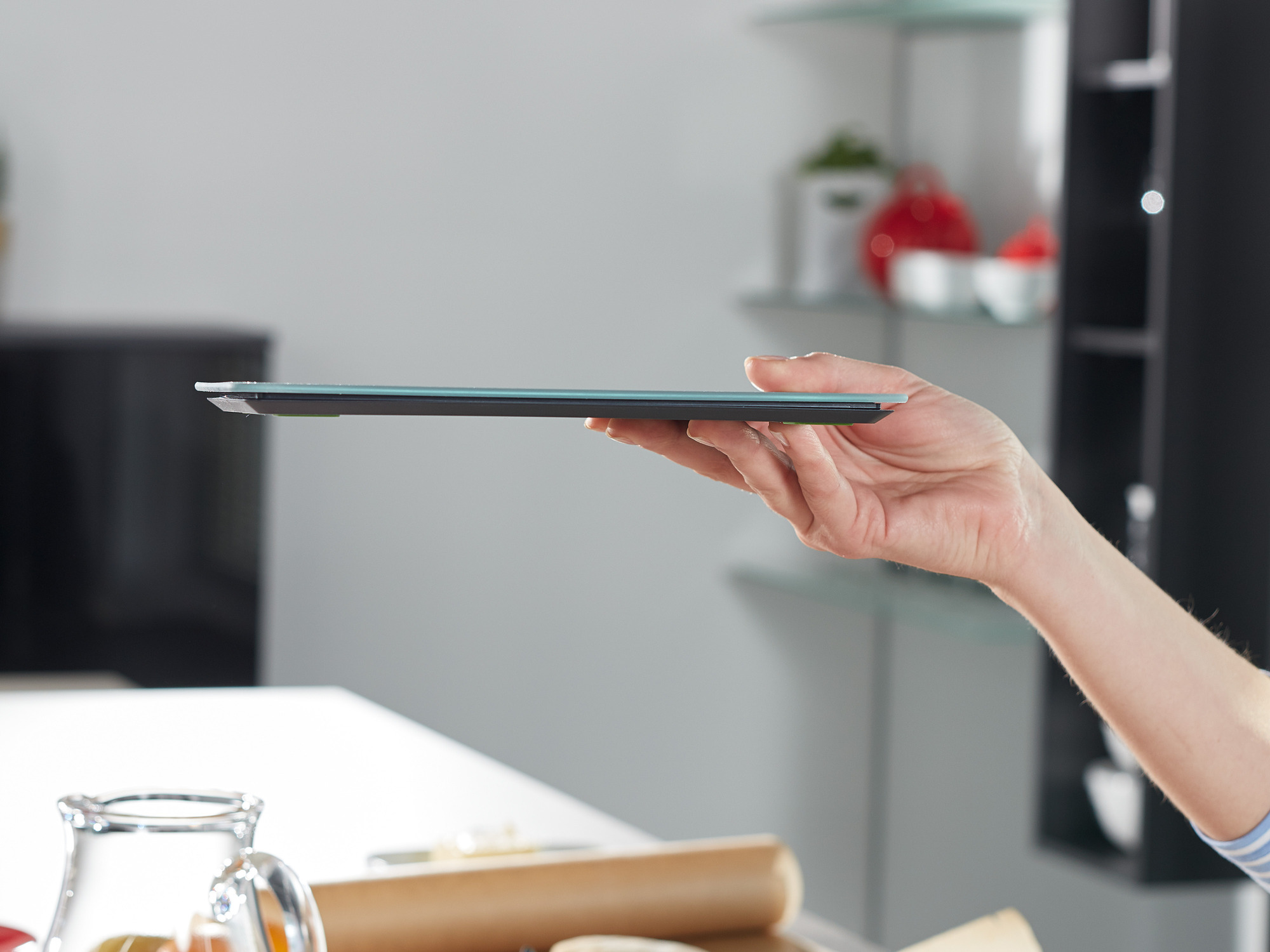 Dłoń trzymająca szklaną deskę do krojenia. Blat kuchenny widoczny w tle.