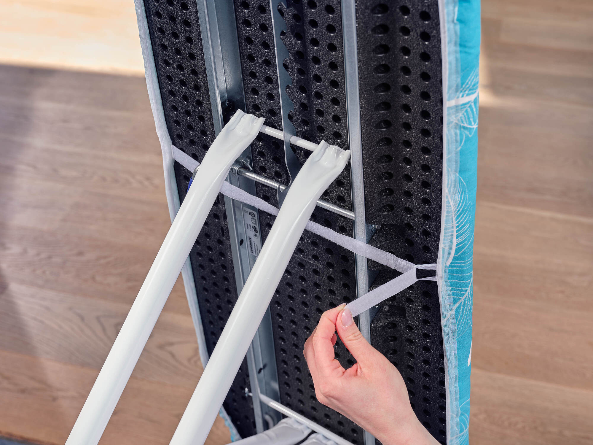 Osoba zabezpiecza pokrowiec deski do prasowania paskami na rzepy, białe nogi i jasna drewniana podłoga.