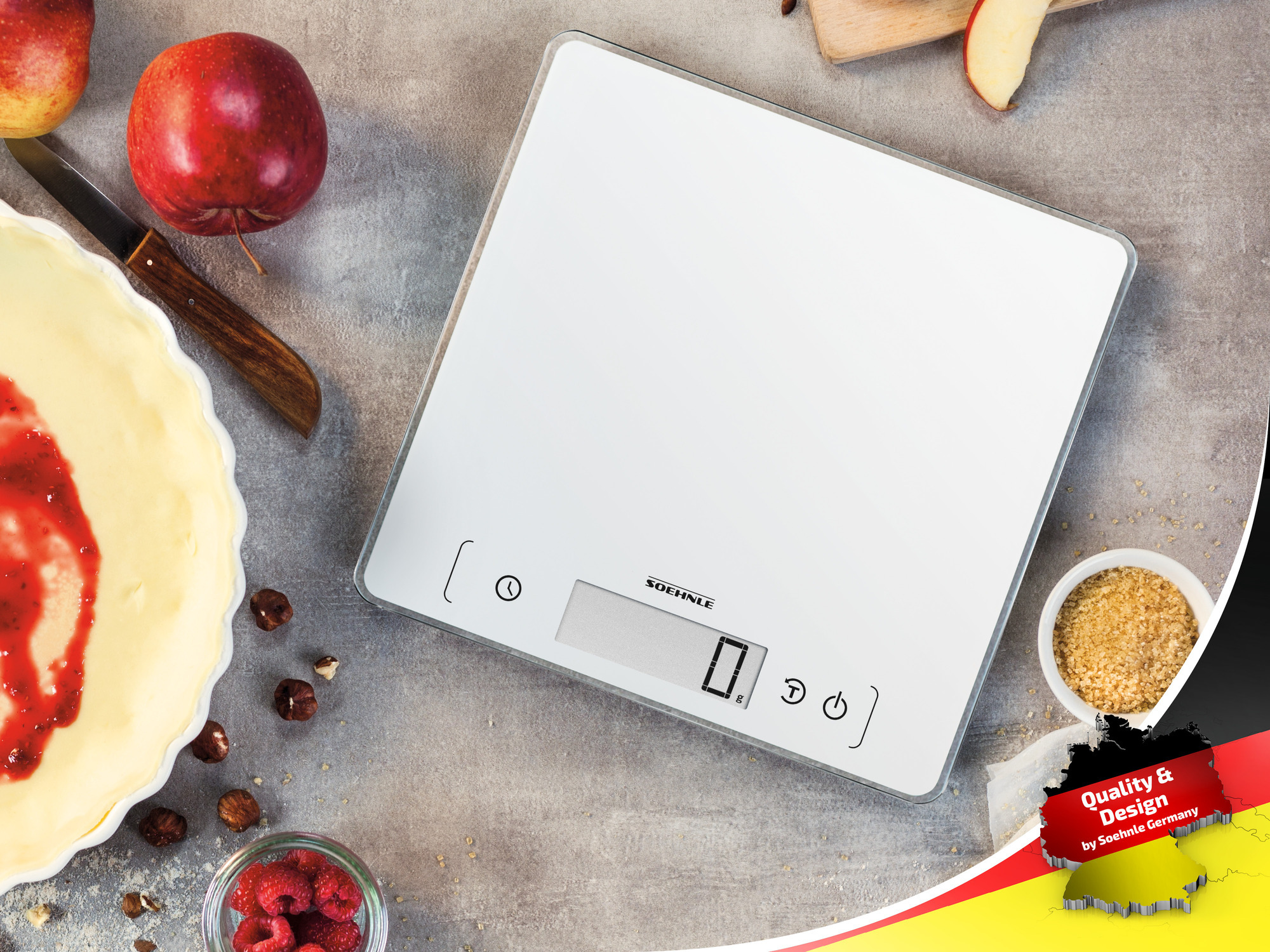 Biała cyfrowa waga kuchenna z otaczającymi ją produktami spożywczymi: jabłkami, ciastem, malinami i orzechami na szarej powierzchni.