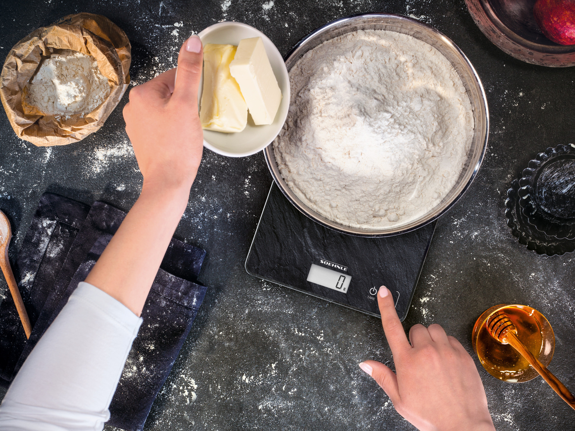 Ręce ważące mąkę i masło na czarnej wadze z innymi składnikami.