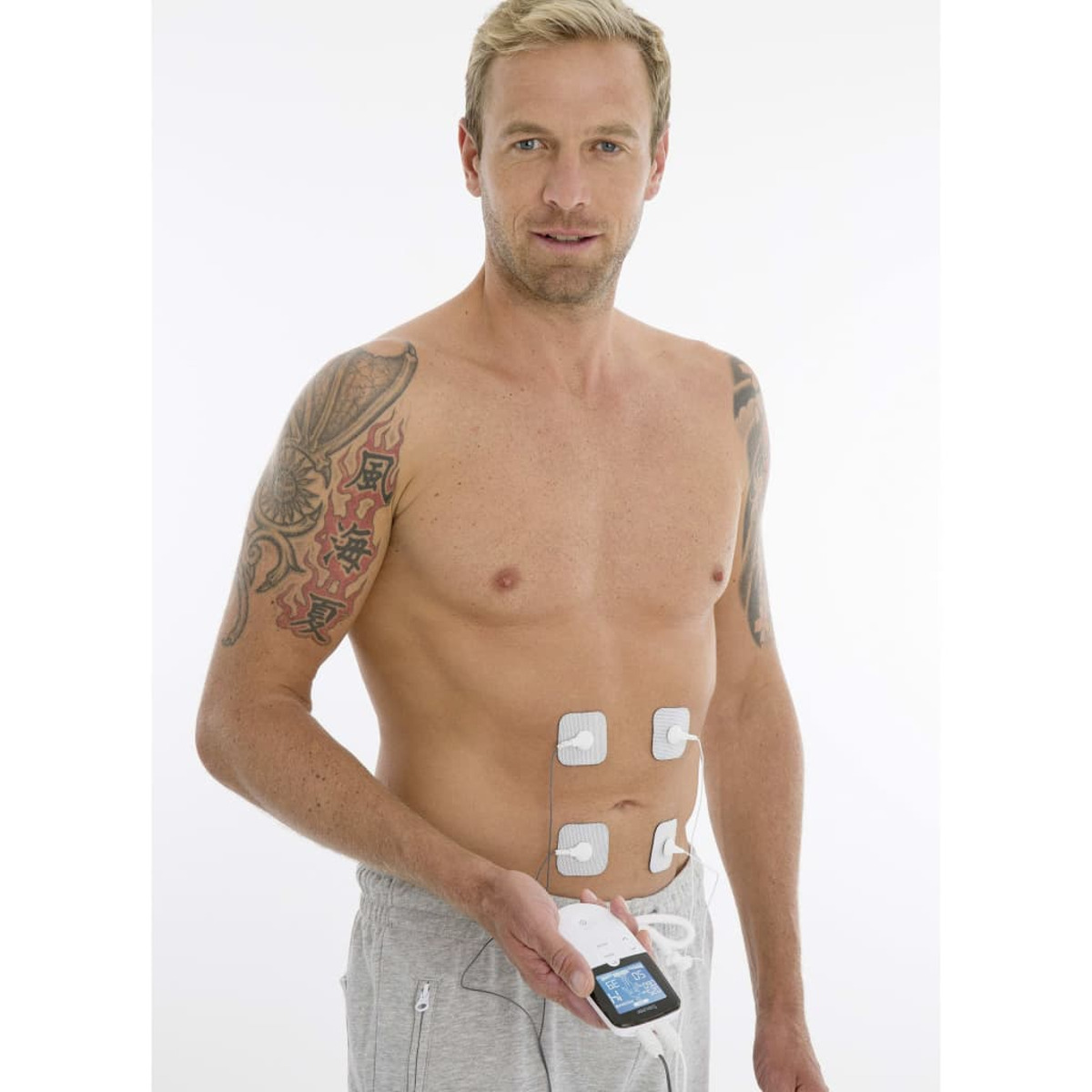 Ein Mann mit Tätowierung am linken Arm. Er hat Elektroden an der Brust und hält ein Gerät.