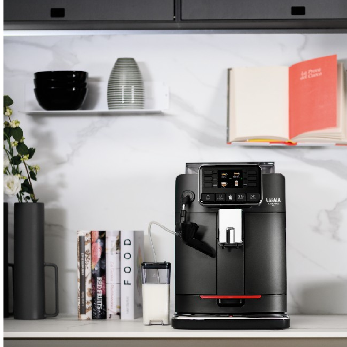 Eine schwarze Espressomaschine auf einer Theke mit einem Glasmilchbehälter. Bücher und Dekorationen sind im Hintergrund.