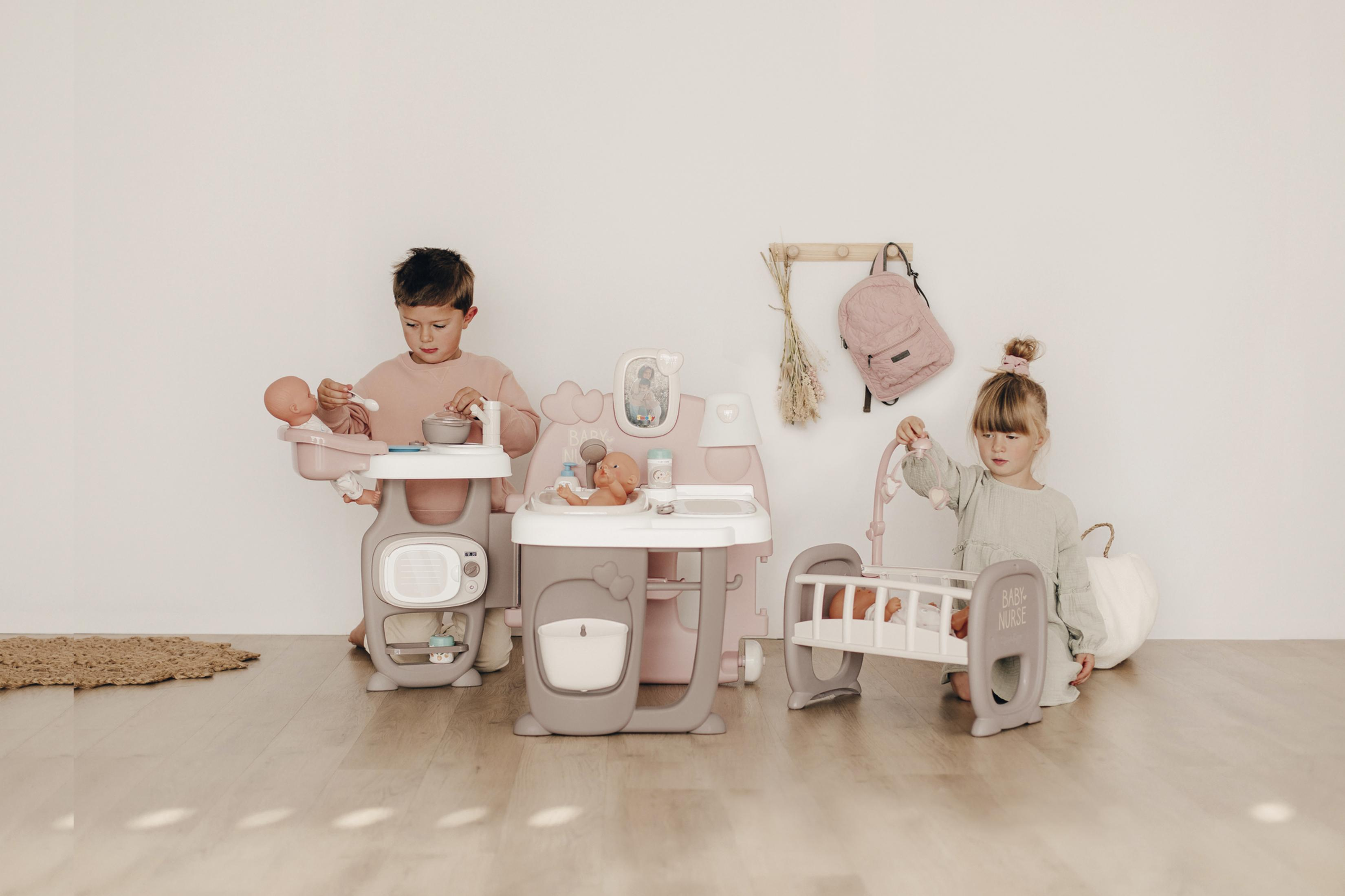 Zwei Kinder spielen mit einer Spielzeugküche und Babypuppen. Im Bild ist ein Kinderbett zu sehen.