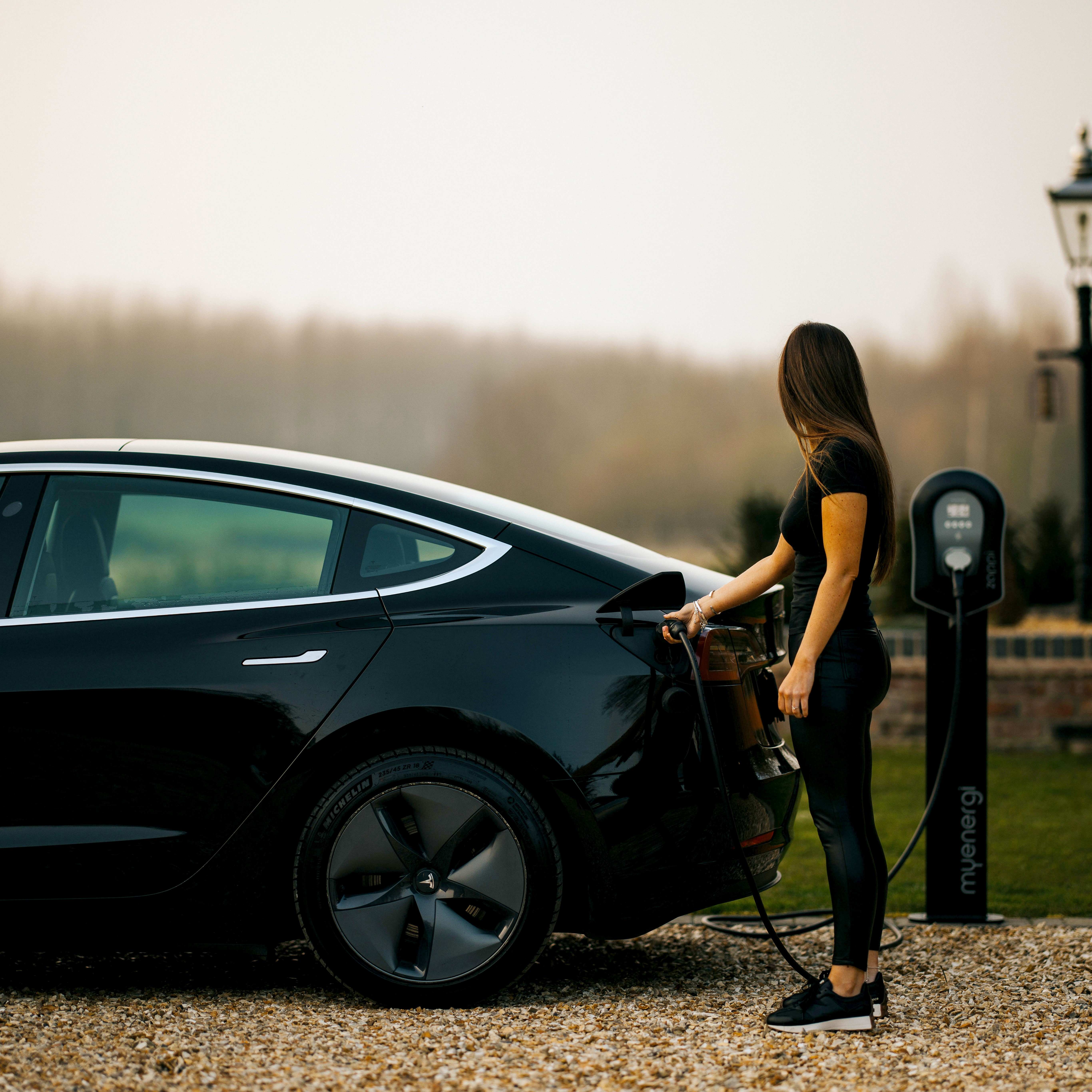 Donna che carica un'auto elettrica nera presso una stazione di ricarica all'aperto.