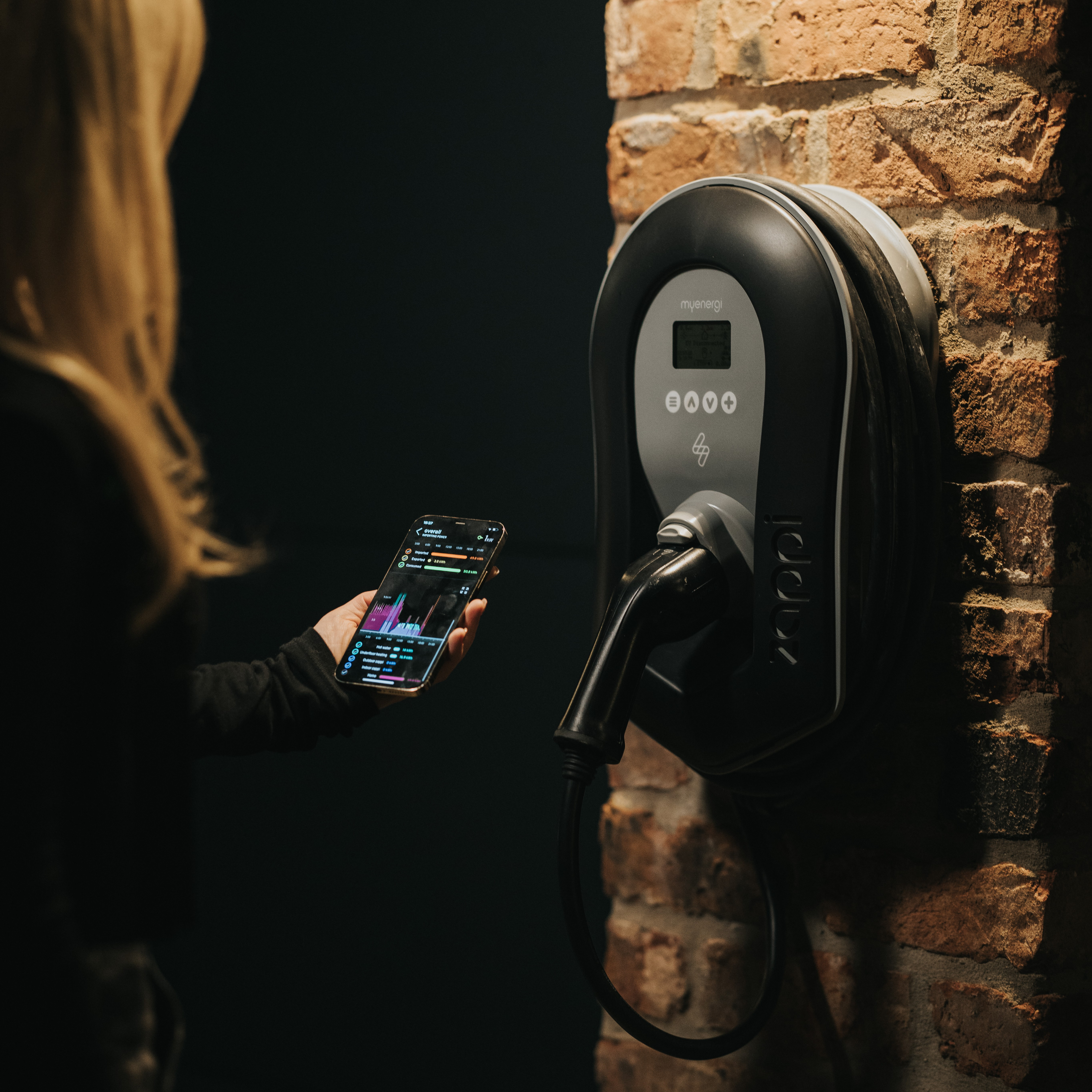 A person holds a phone, viewing a graph, near a charging station mounted on a brick wall.