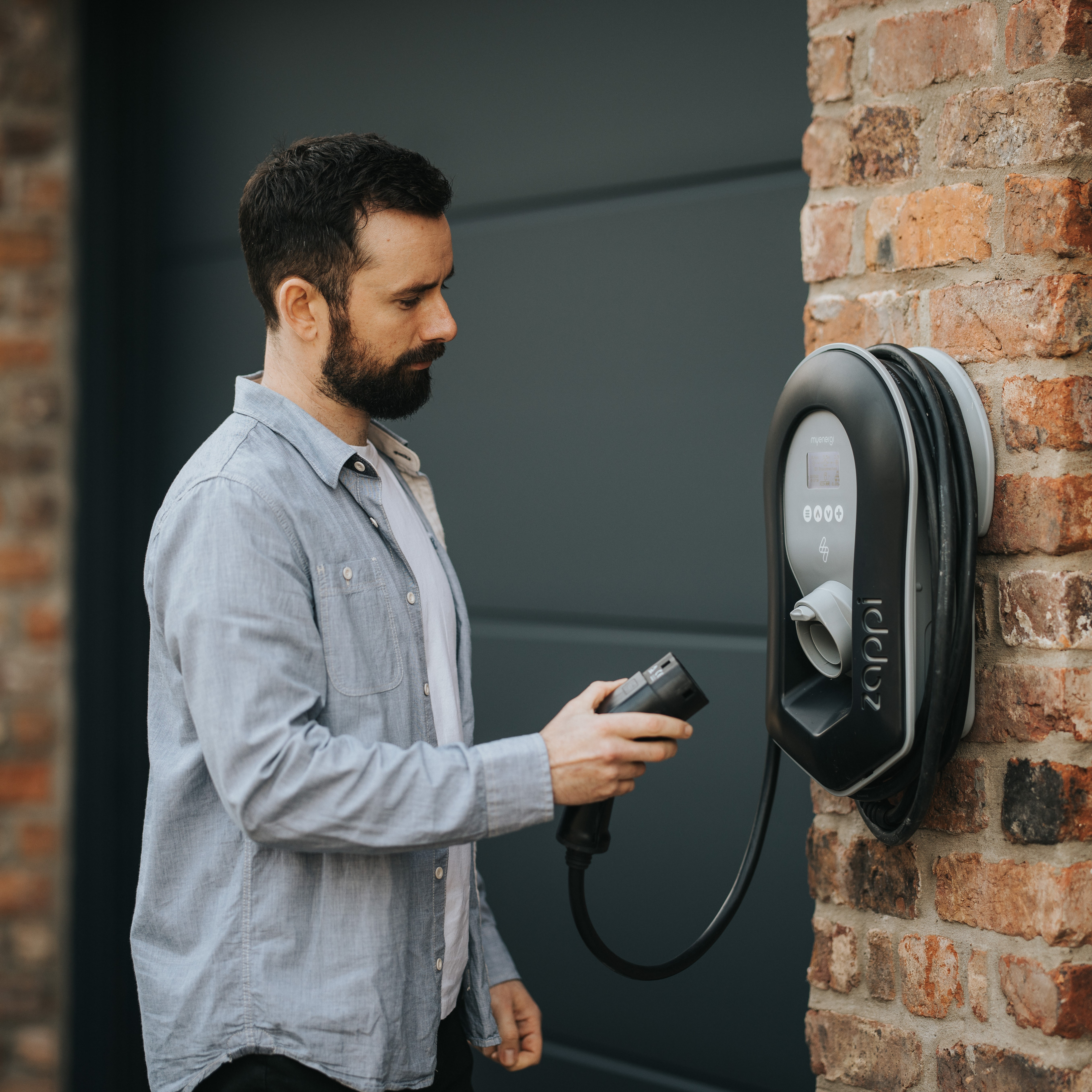 Man with beard charging an EV with a charging station on a brick wall, gray door behind.